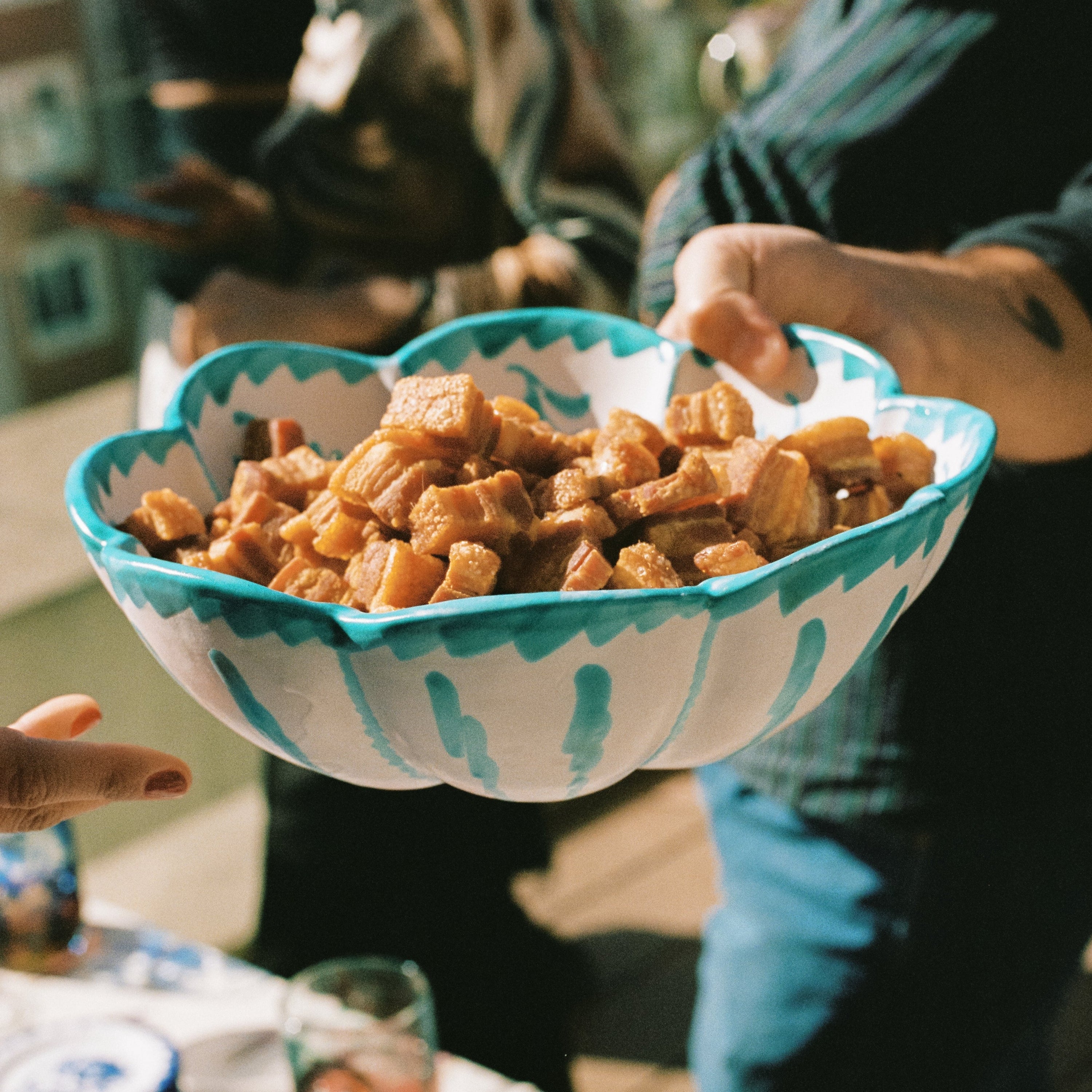 Handmade turquoise leaf-shaped serving bowl filled with food and held during a shared dinner gathering, featuring hand-painted ceramic details inspired by Mediterranean tableware traditions. - lifestyle