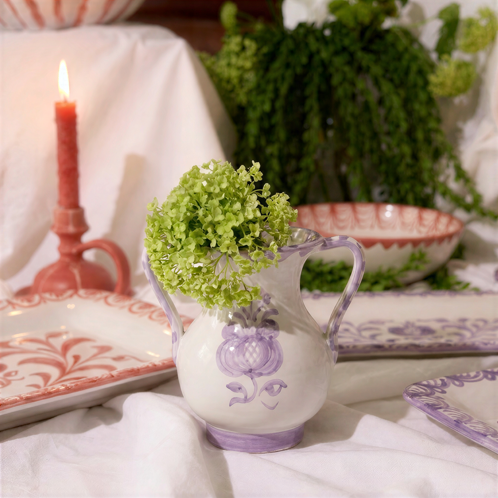 A white ceramic pitcher vase with purple floral motifs and a green hydrangea stem, styled on a white draped tablecloth with a red candle, patterned plates, and green foliage in the background. - lifestyle