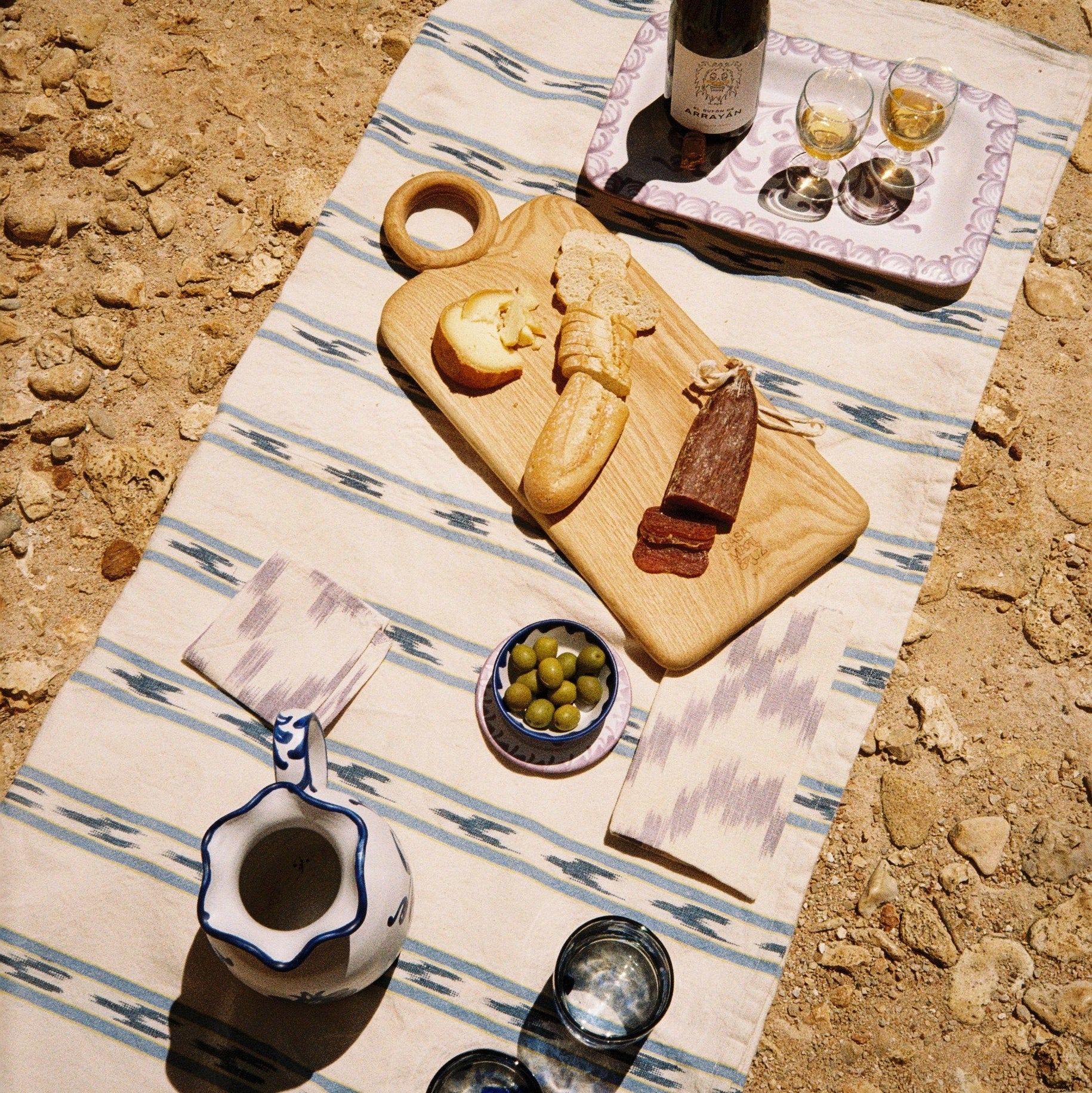 Outdoor setting with a wooden cutting board, wine, and snacks on a striped tablecloth.