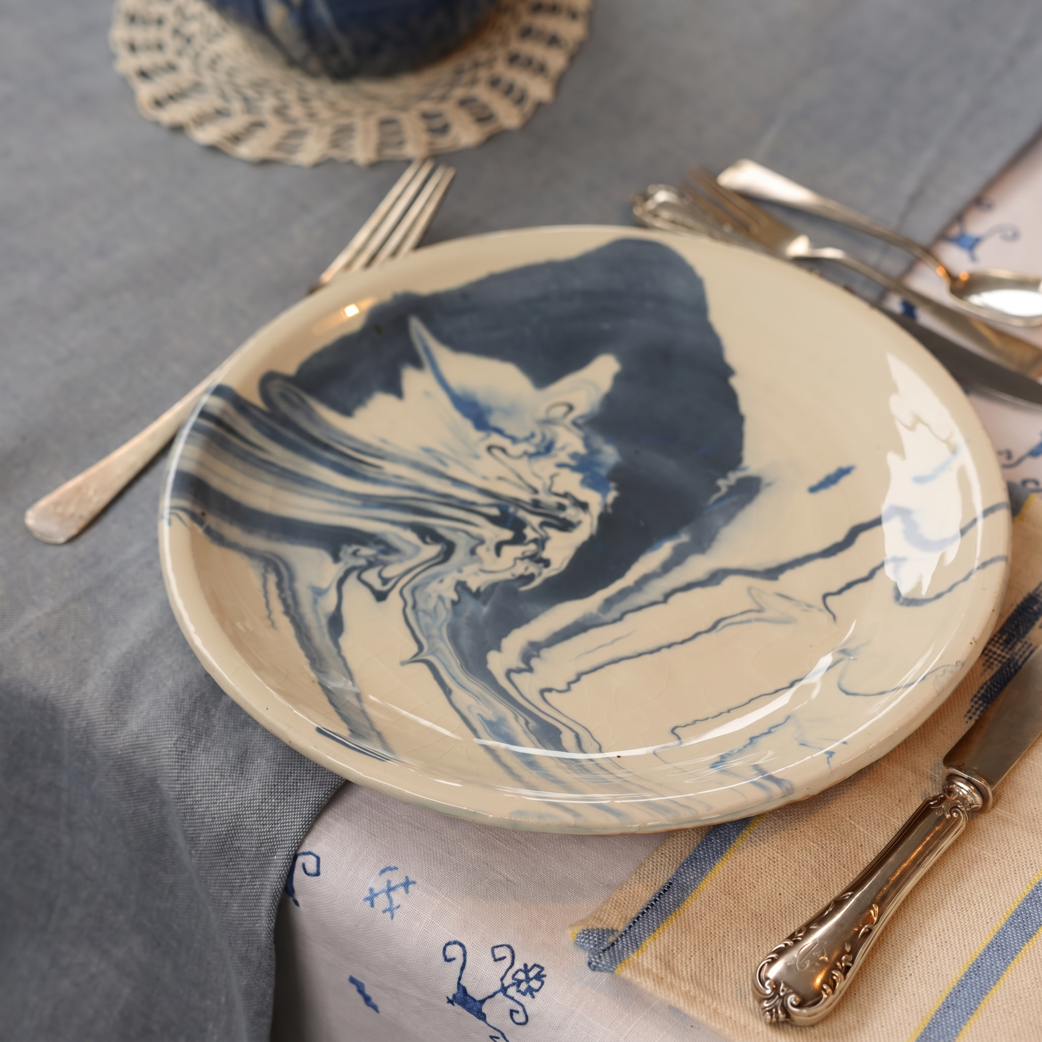 A round ceramic plate with a cream base and a dark blue marbled abstract pattern, styled on a gray tablecloth with silver cutlery and a patterned napkin. - lifestyle