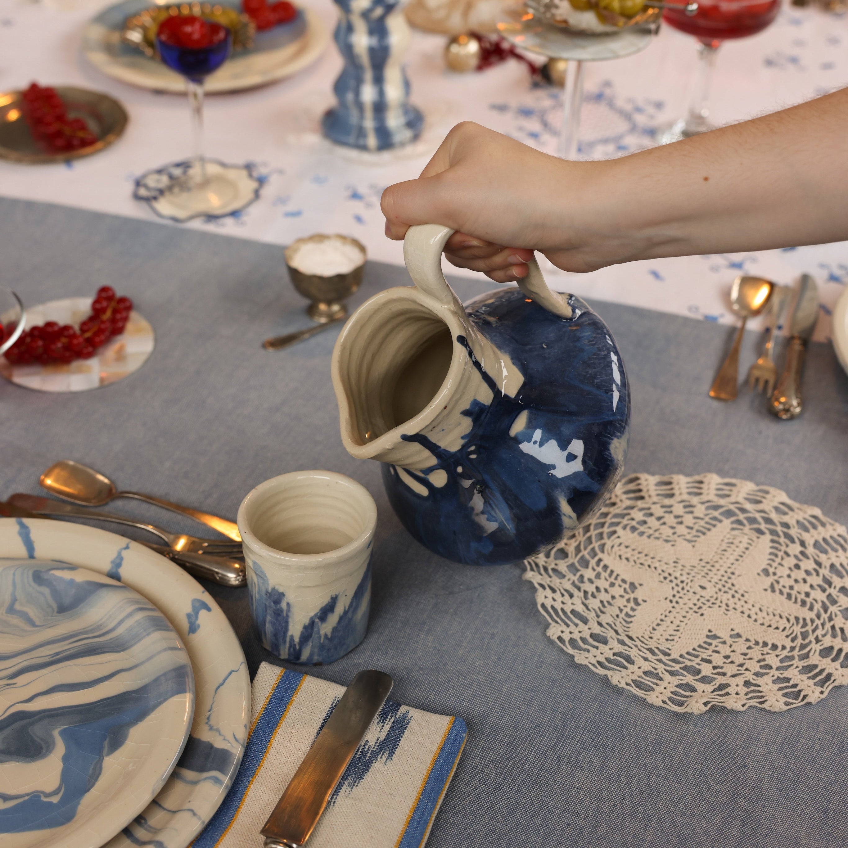Person pouring from a blue and white pitcher on a decorated table with plates, cutlery, and glasses.