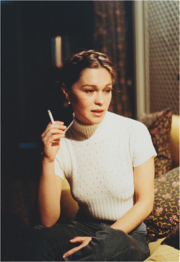 Woman sitting on a couch holding a cigarette in a dimly lit room.