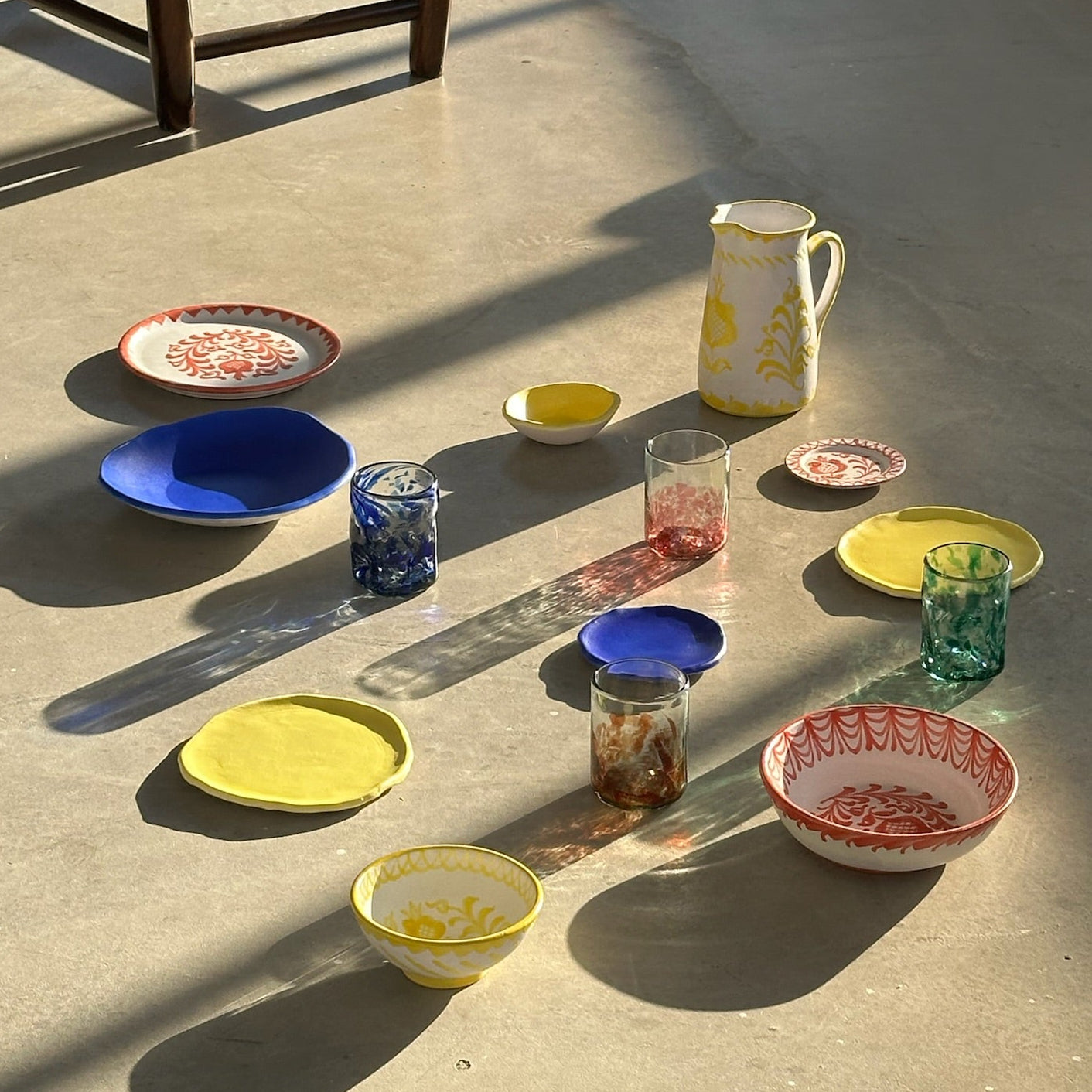 Colorful ceramic dishes and glasses arranged on a concrete floor with a brown leather ottoman in the background. - lifestyle