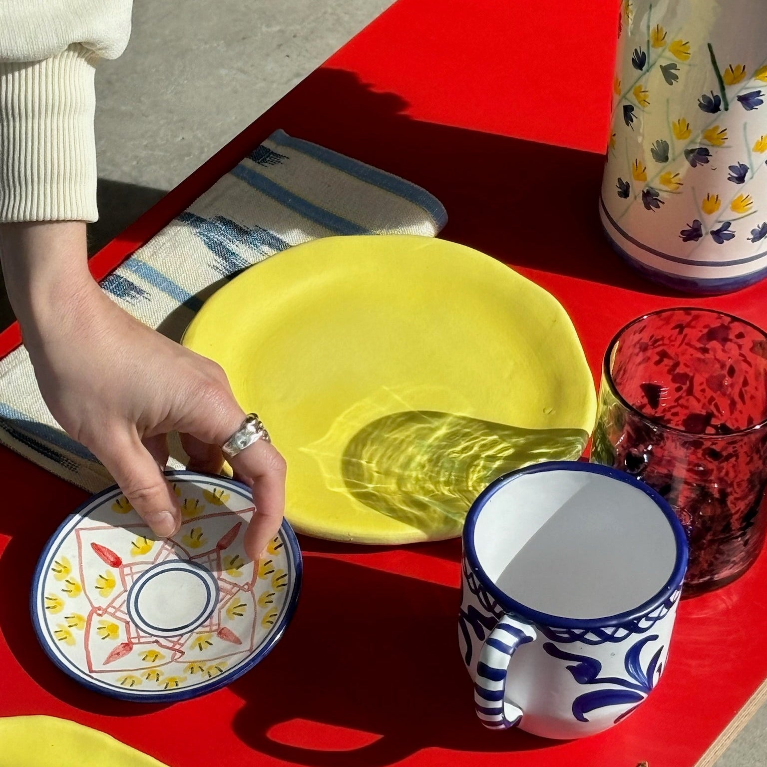Person holding a small decorative plate on a red table with colorful ceramic items. - lifestyle