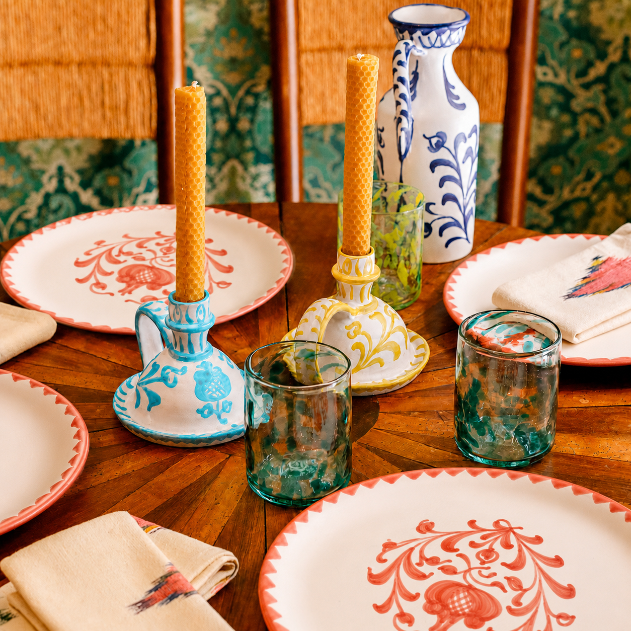 A styled tabletop scene with decorative ceramic plates, patterned napkins, two candlesticks with yellow candles, and two teal glass tumblers on a wooden table. - lifestyle