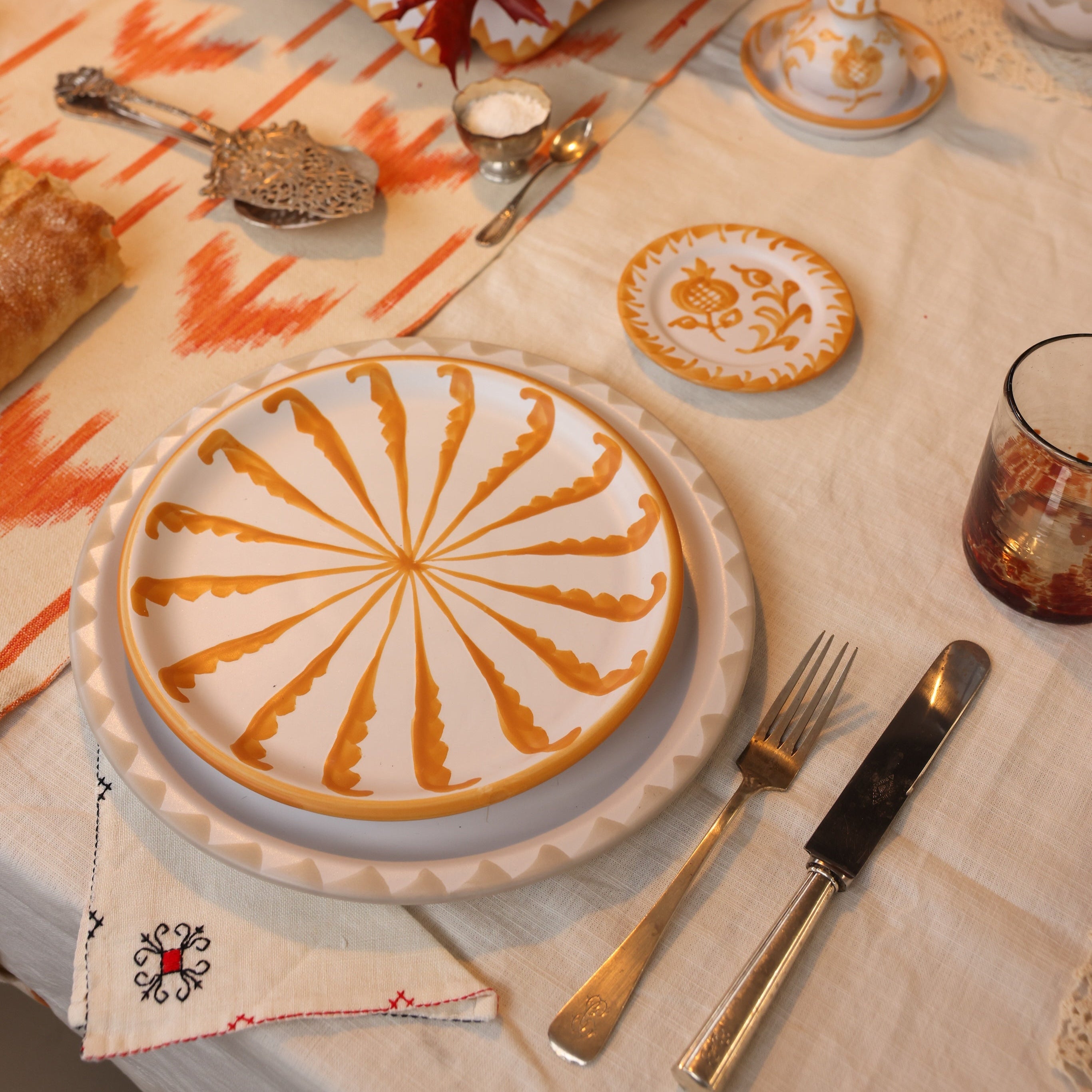 Dining table setting with decorative plates, cutlery, and a glass on a patterned tablecloth.