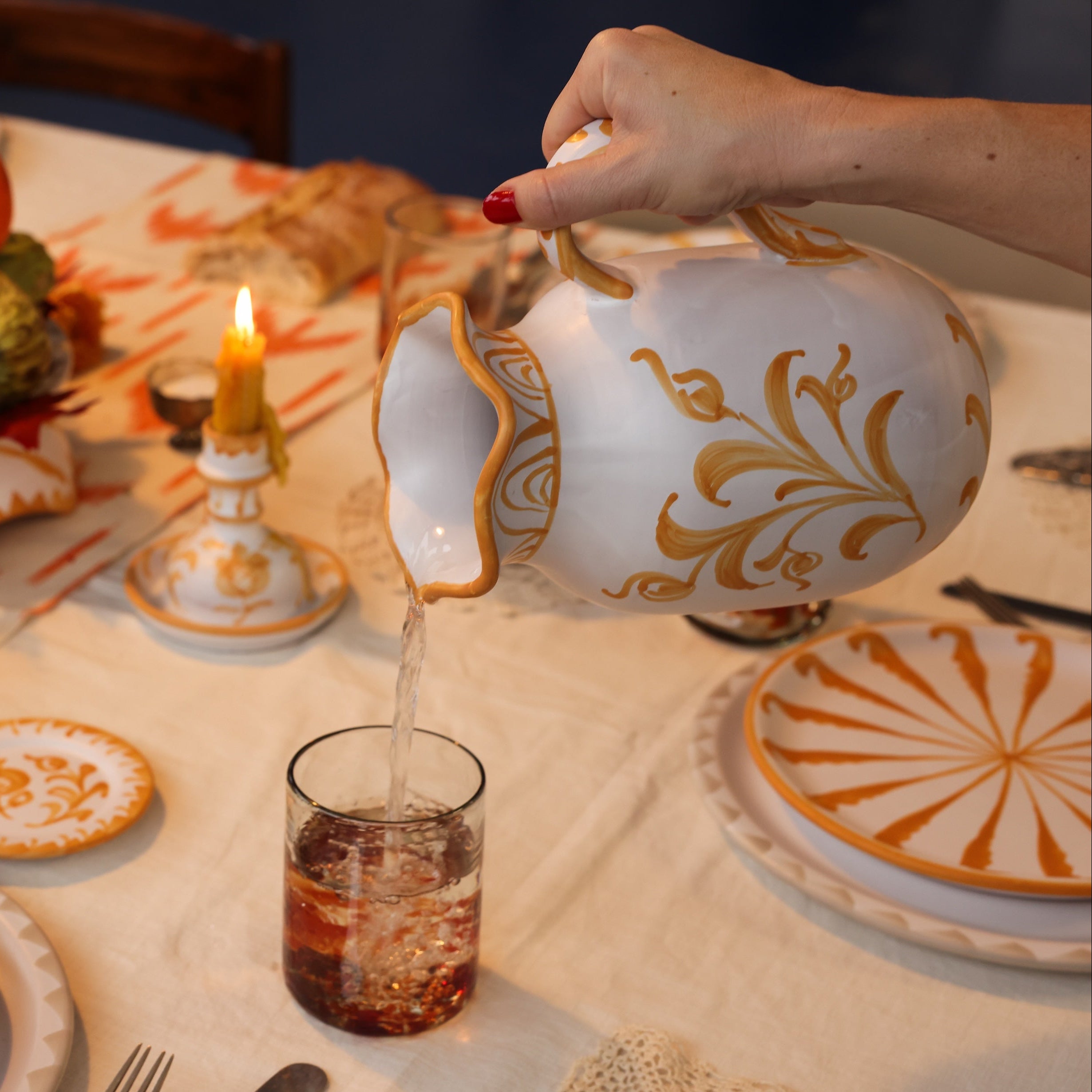 Person pouring liquid from a decorative teapot into a glass on a table set with plates and candles.
