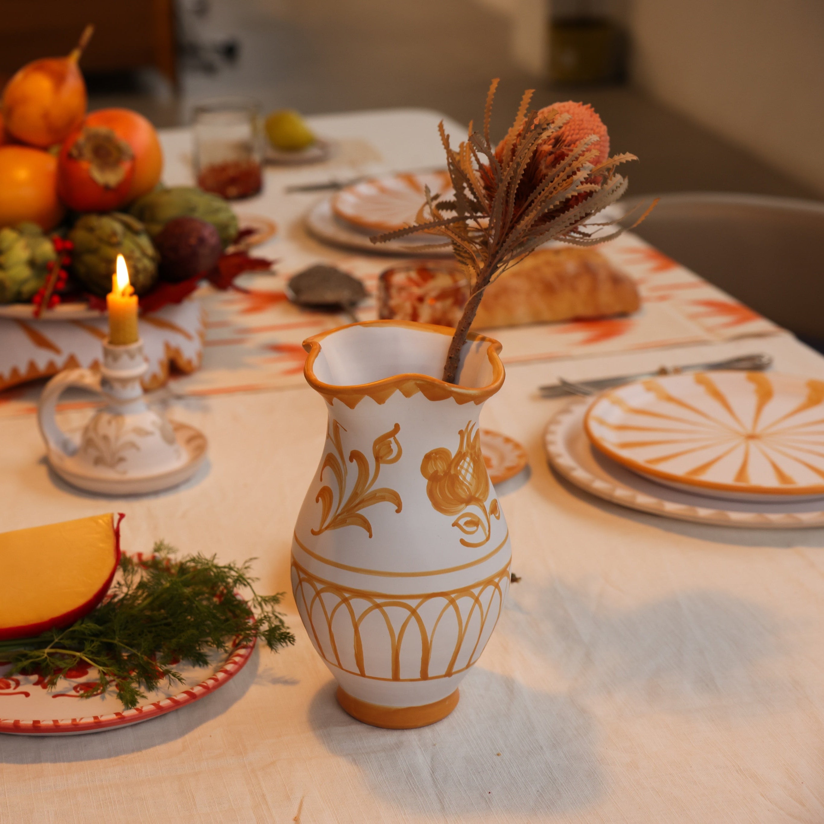 Decorative table setting with a vase, fruit, and candles on a tablecloth.