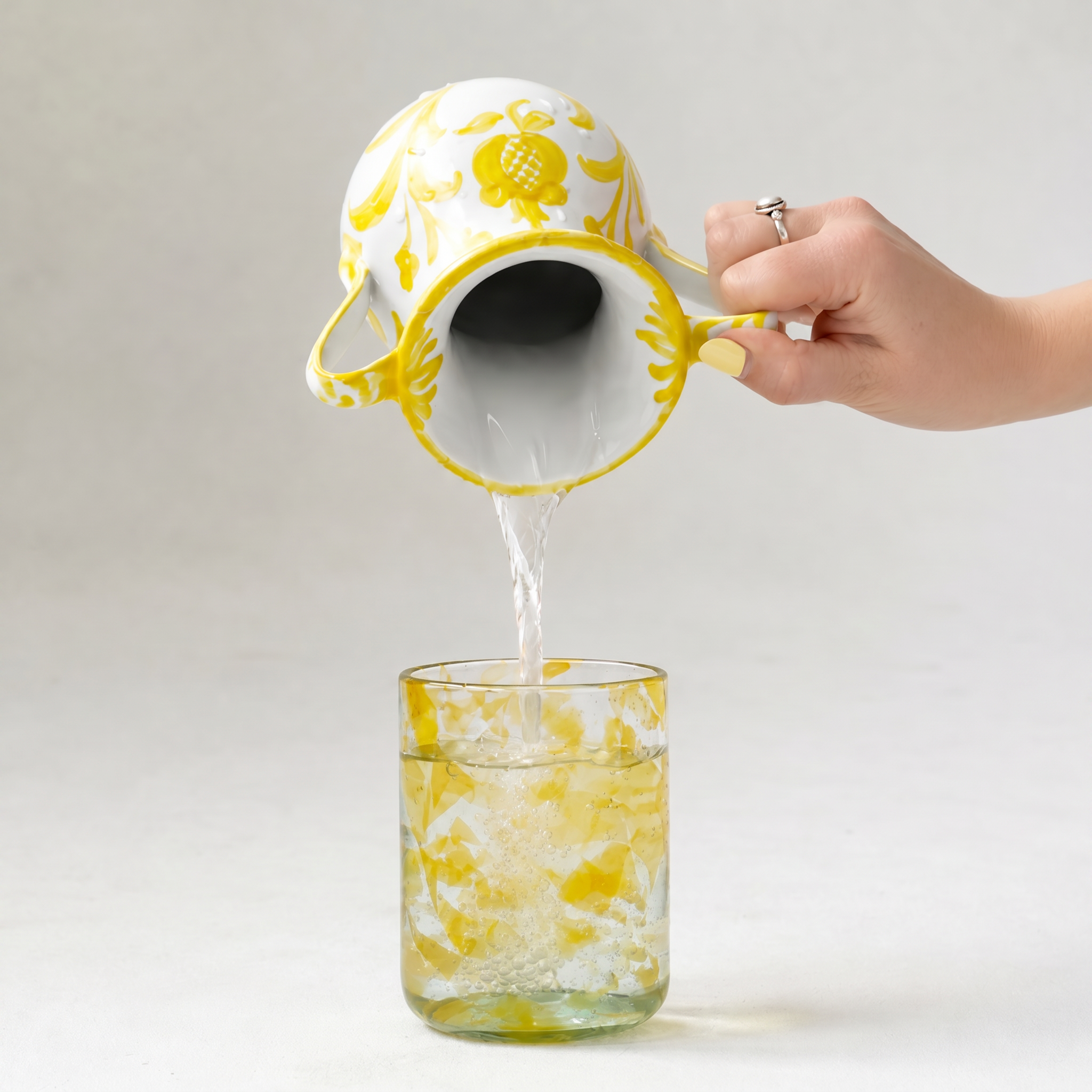 A hand with yellow nail polish holds a white ceramic pitcher with a yellow floral pattern, pouring water into a clear glass tumbler with a yellow marbled pattern, against a light gray background. - lifestyle