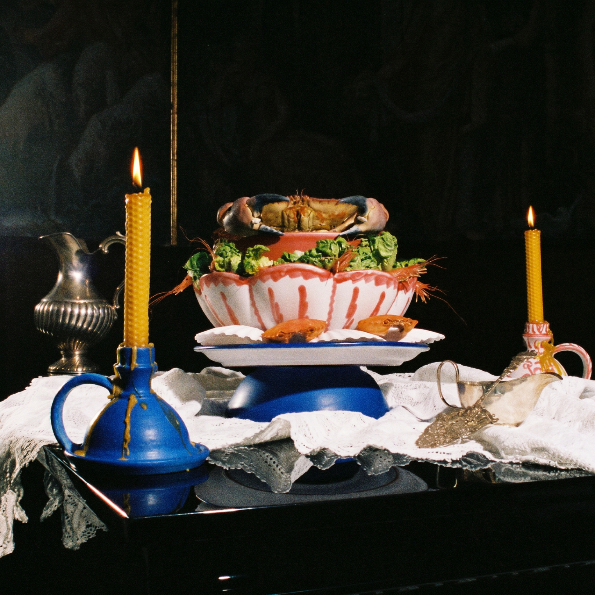 A still life tabletop scene with a decorative blue and white floral serving dish holding shrimp and broccoli, flanked by two lit yellow candles in blue and pink holders, with a silver pitcher and white cloth against a dark background. - lifestyle