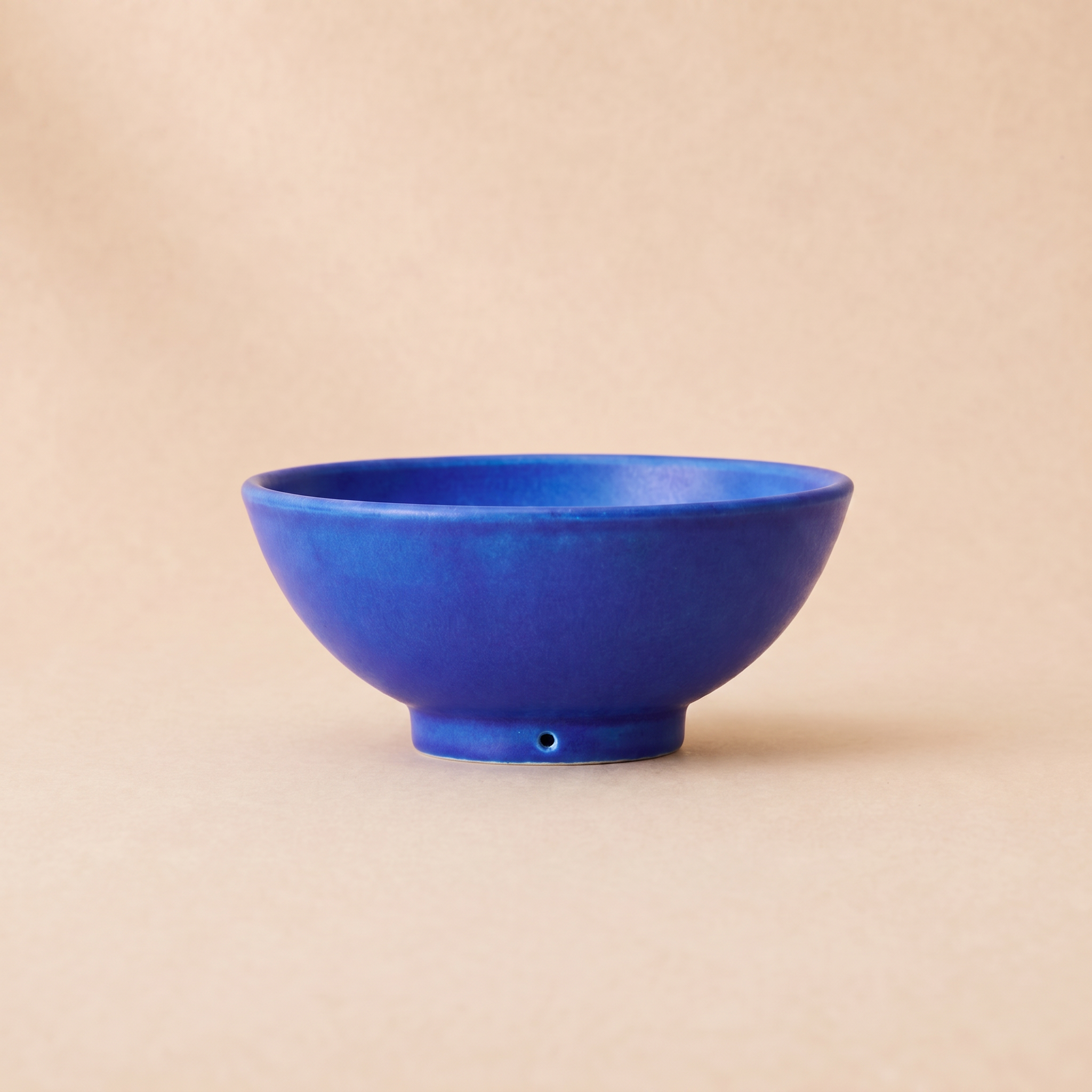 A small ceramic bowl in a rich cobalt blue glaze, set against a solid beige background.