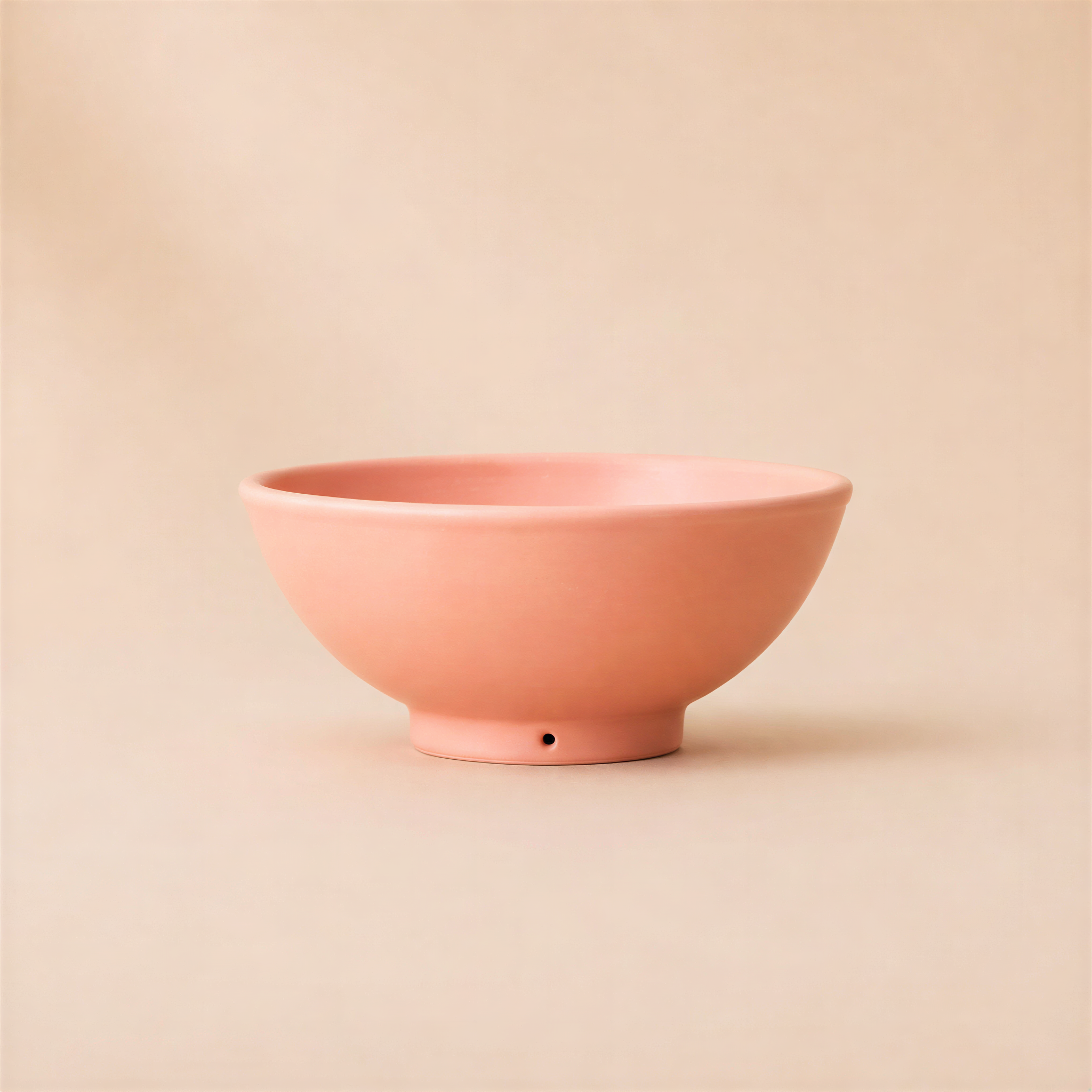 A minimalist dusty coral ceramic bowl with a small drainage hole at the base, set against a solid, warm beige background.