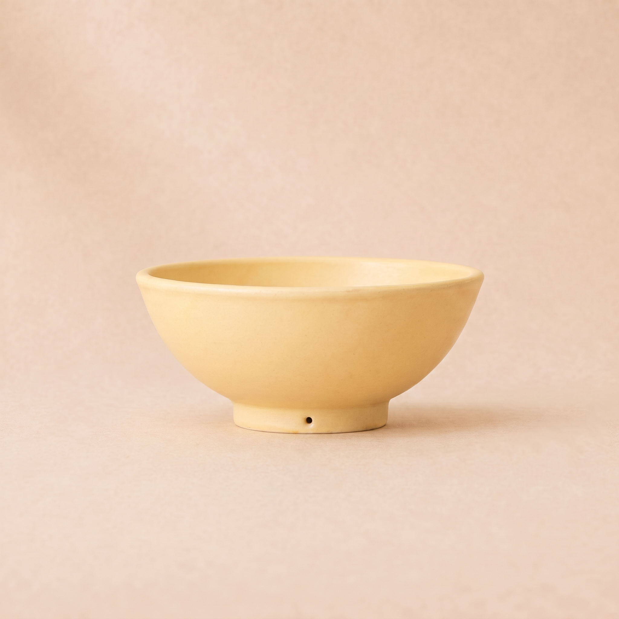 A minimalist ceramic bowl in a pale pastel yellow, featuring a small hole on its base, set against a solid, warm beige background.