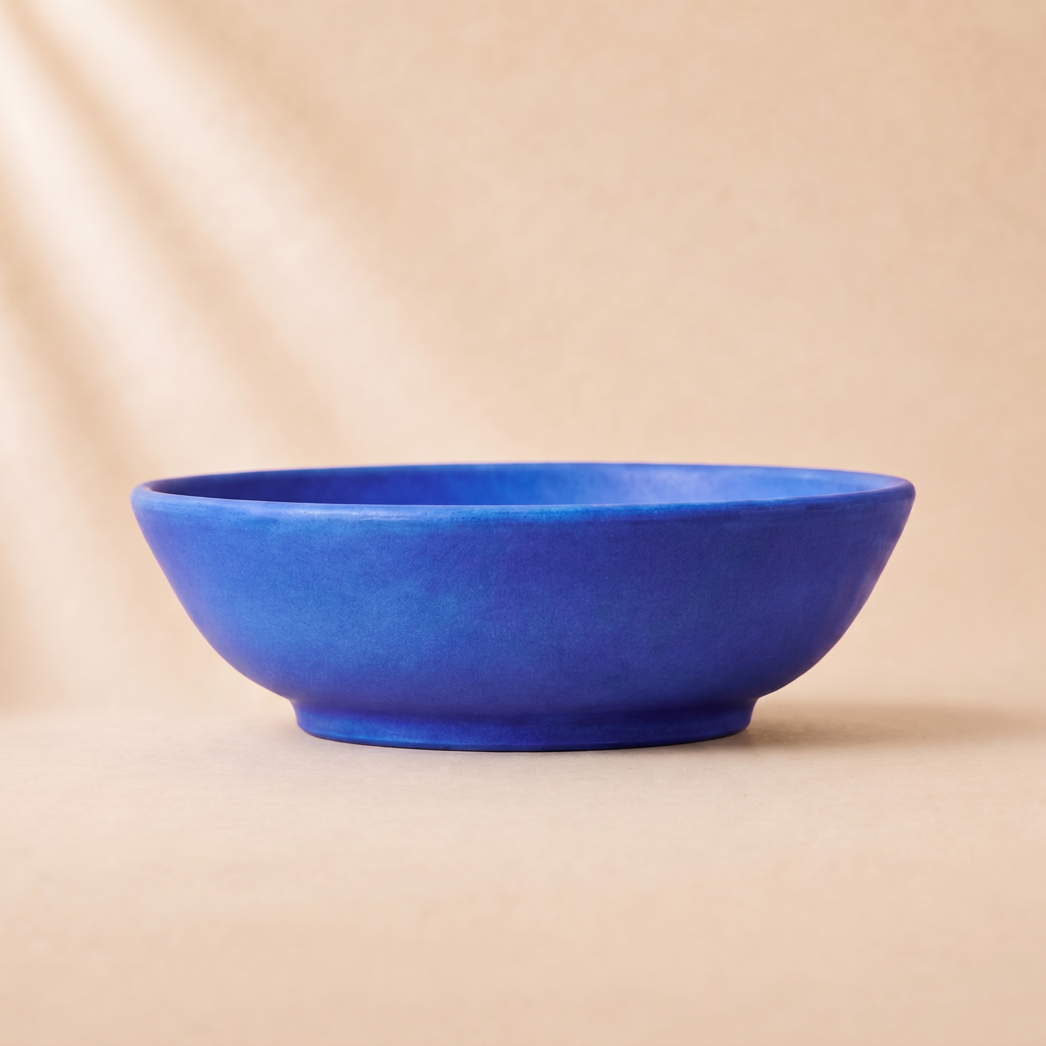 A minimalist terracotta blue ceramic bowl with a matte finish, set against a warm beige background with soft diagonal shadows.