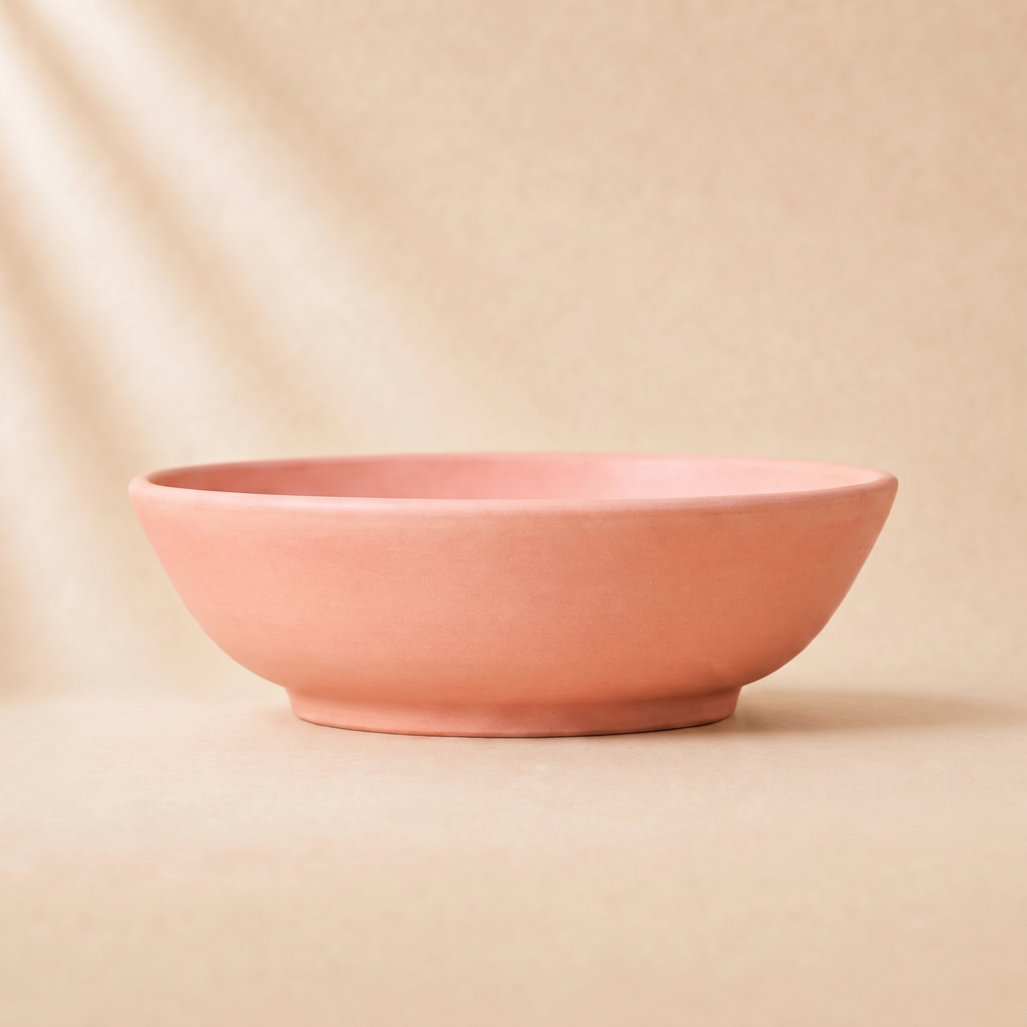 A minimalist terracotta coral ceramic bowl with a matte finish, set against a warm beige background with soft diagonal shadows.