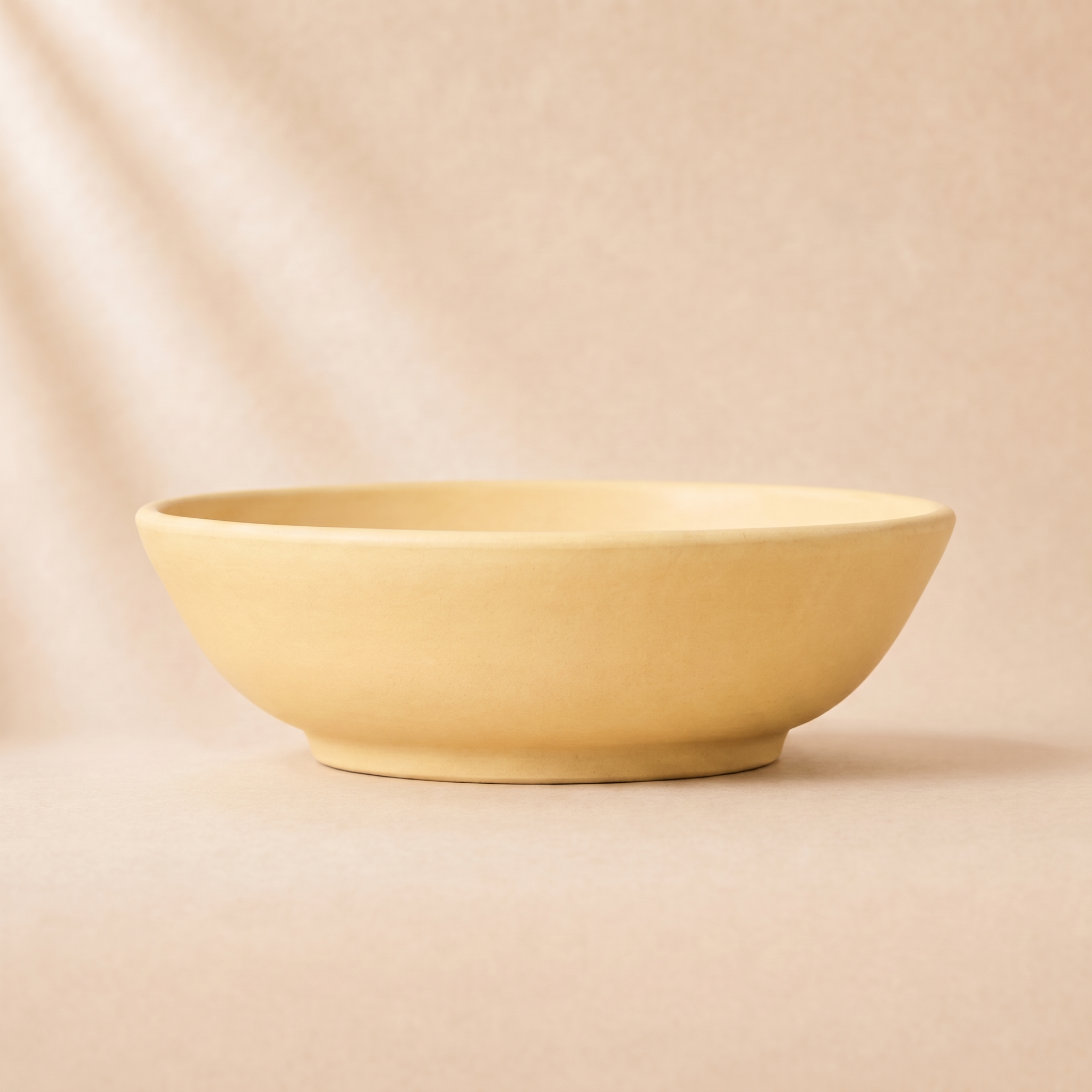 A minimalist, matte ceramic bowl in a pale pastel yellow, photographed on a matching warm beige background with soft, artistic shadows.