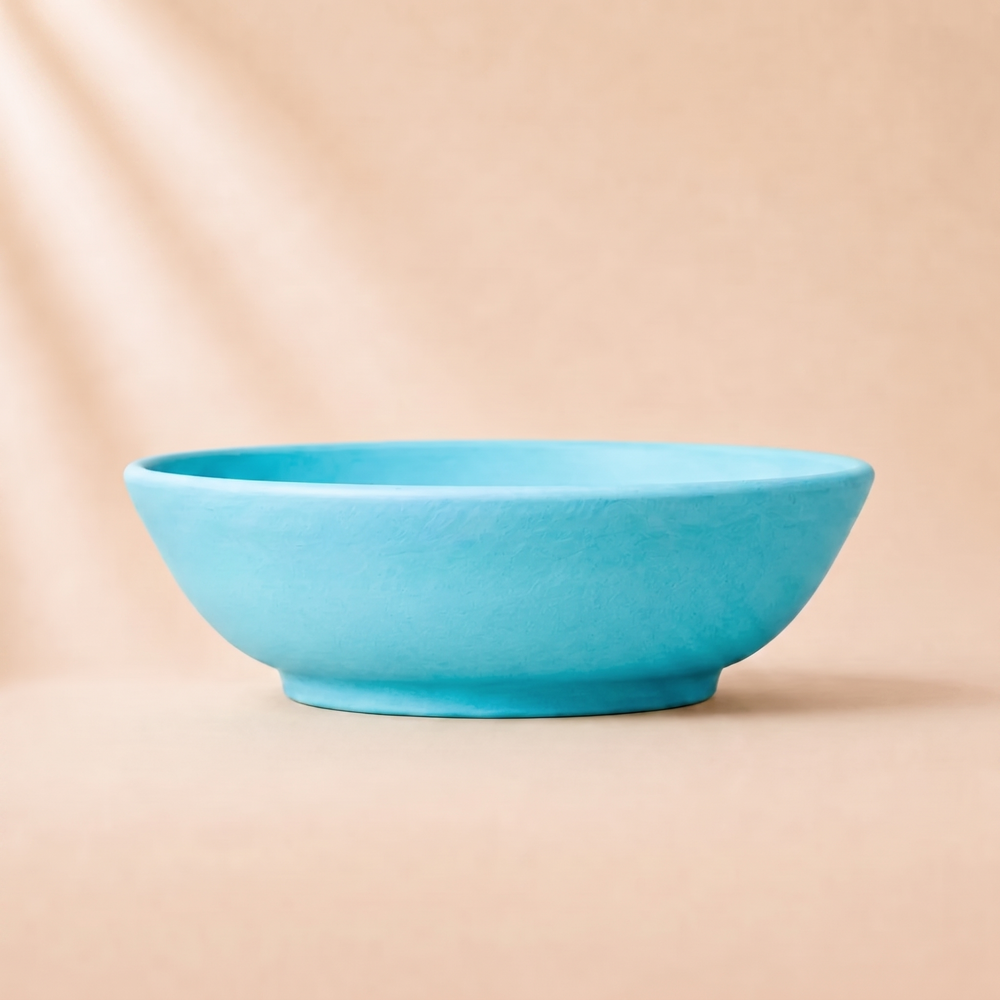 A bright turquoise turquoise ceramic bowl with a matte, textured finish, set against a warm beige background with soft, diagonal light rays.