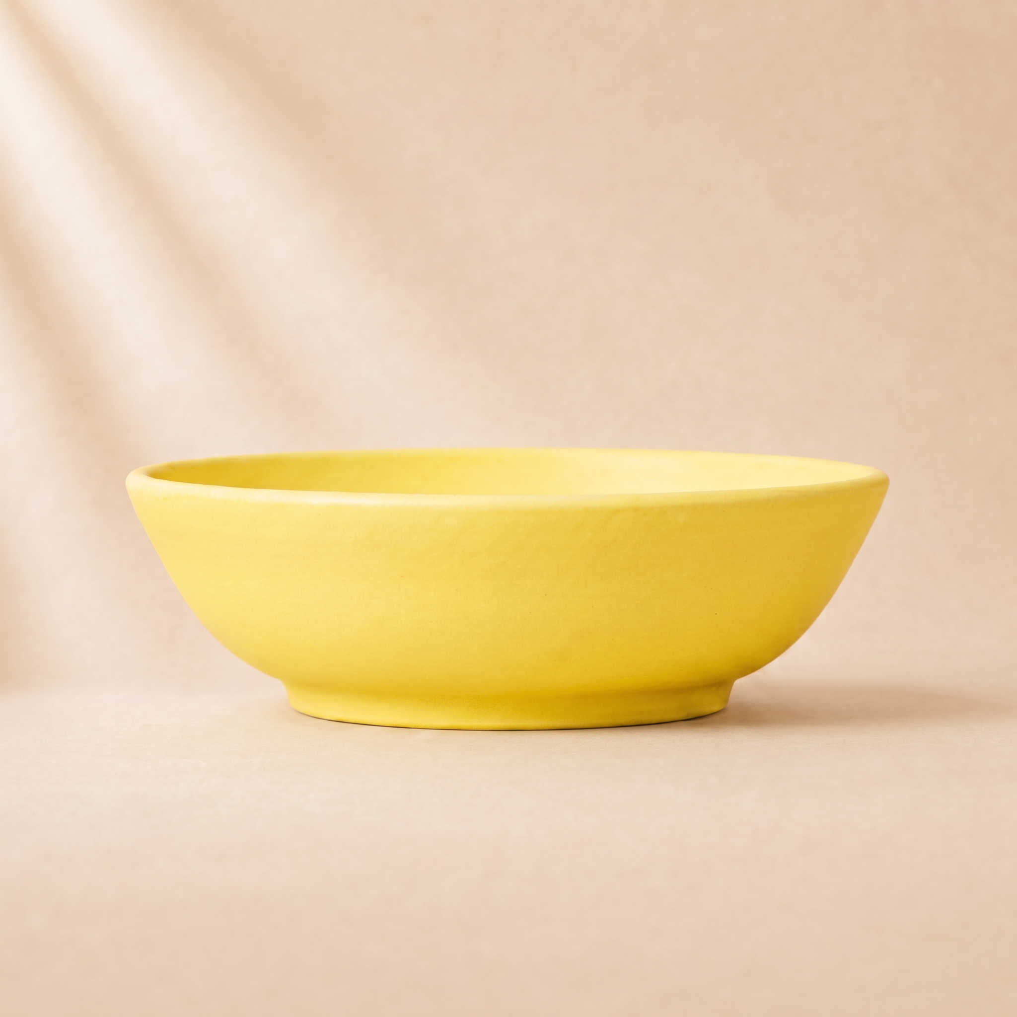 A minimalist terracotta yellow ceramic bowl with a matte finish, set against a warm beige background with soft diagonal shadows.