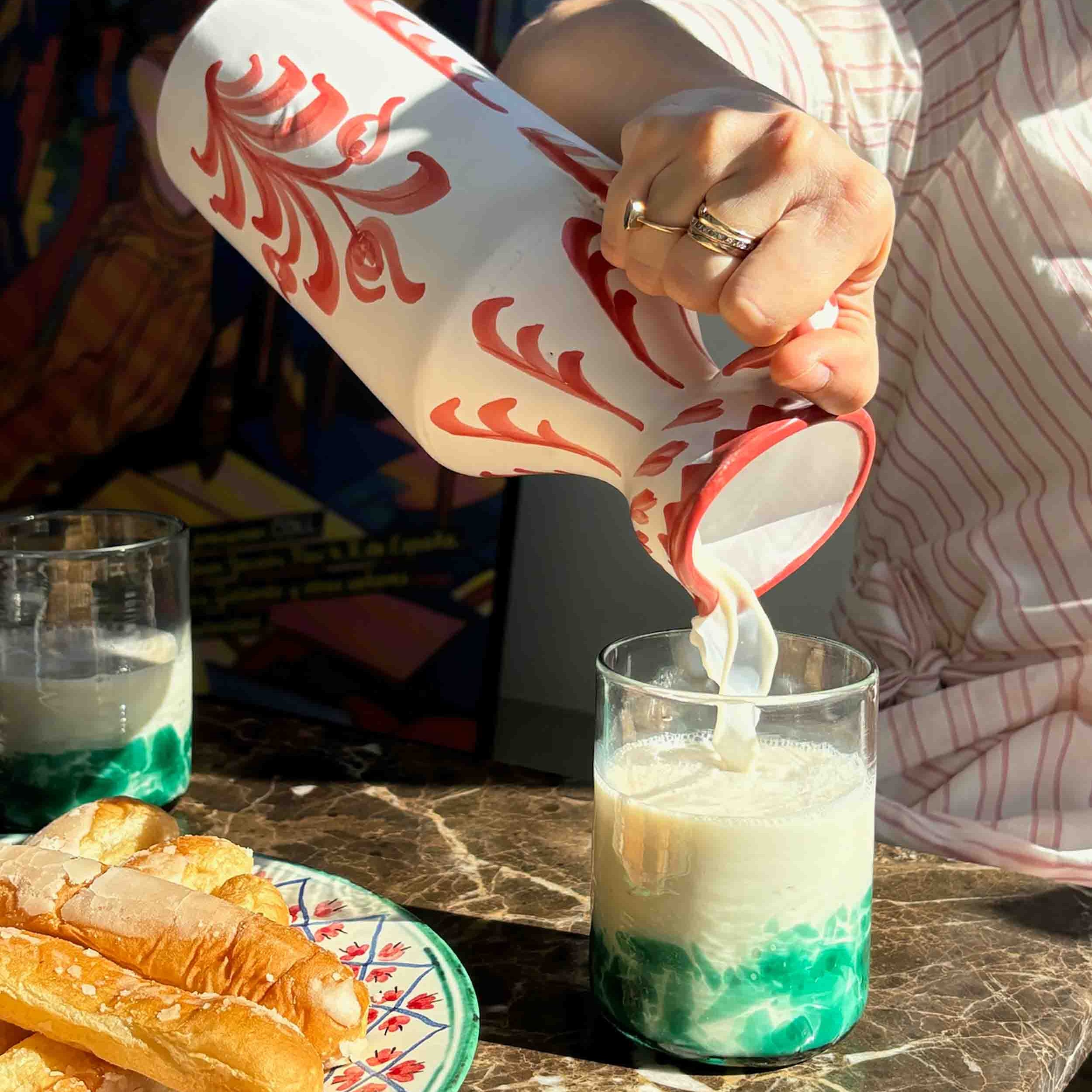 Person pouring a green and white drink from a decorative bottle into a glass on a table with pastries - lifestyle