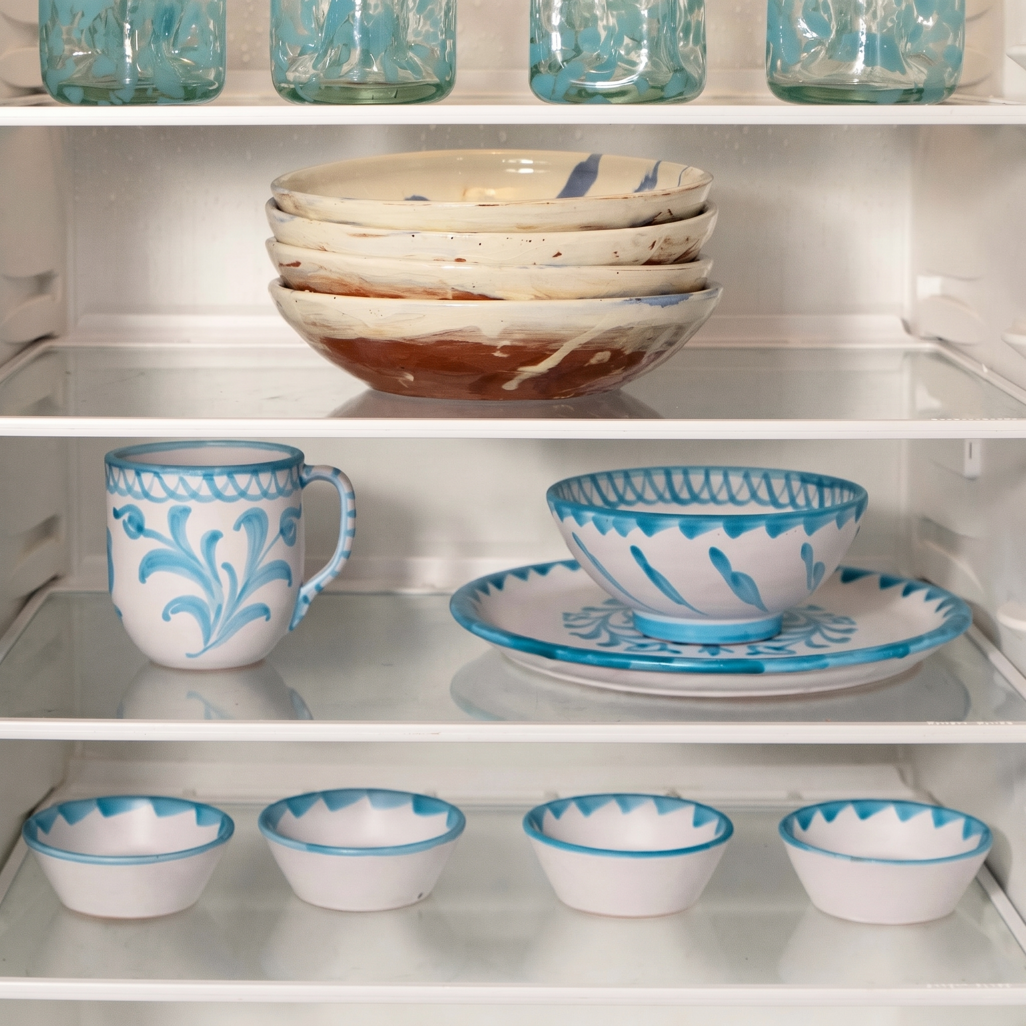 A set of white ceramic tableware with blue patterns, including bowls, plates, cups, and mugs, neatly arranged on glass shelves inside a white refrigerator. - lifestyle