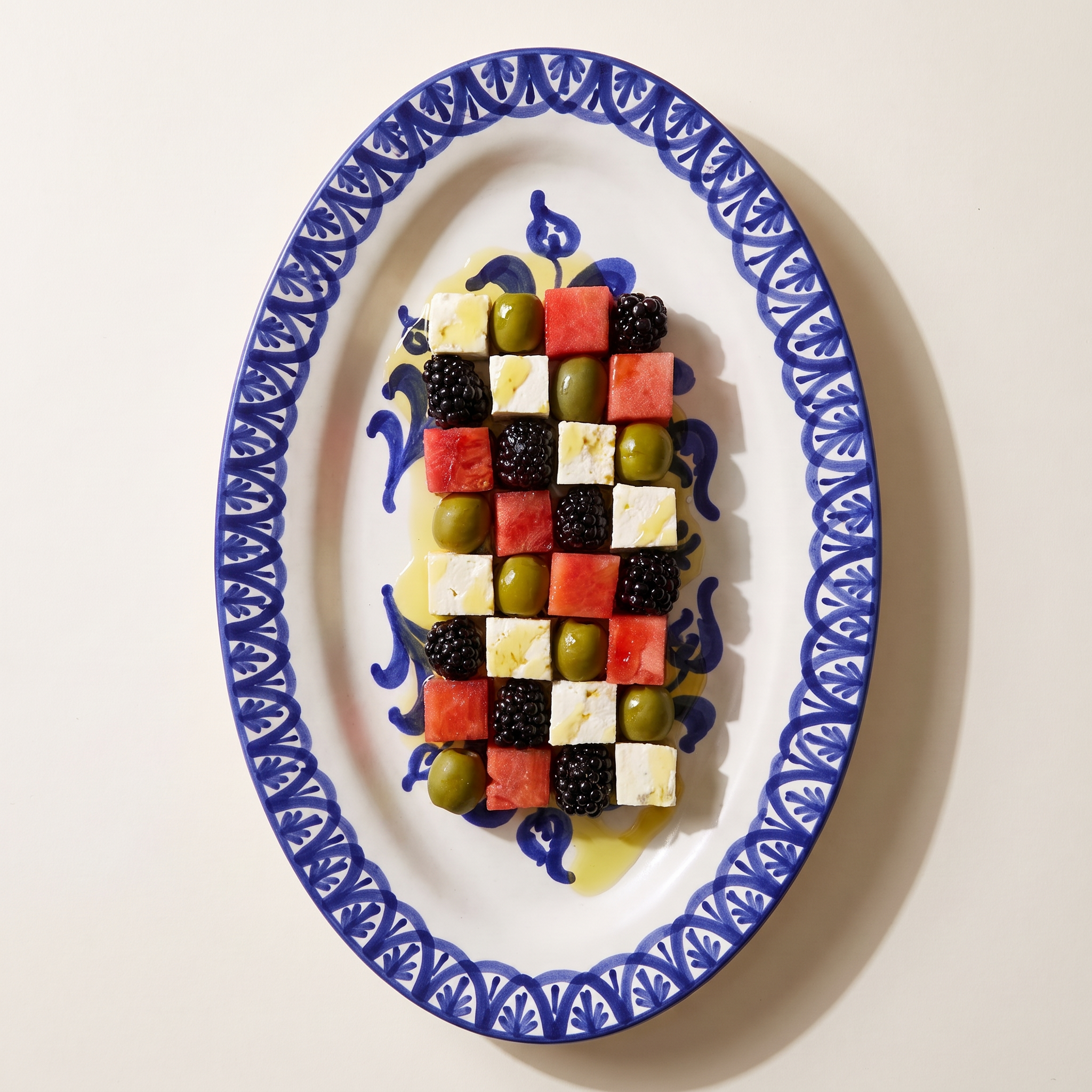 An oval white ceramic platter with a blue patterned border, holding a small appetizing fruit and cheese board arranged in a checkerboard pattern with watermelon, blackberries, olives, and white cheese cubes, on a light beige background. -  lifestyle