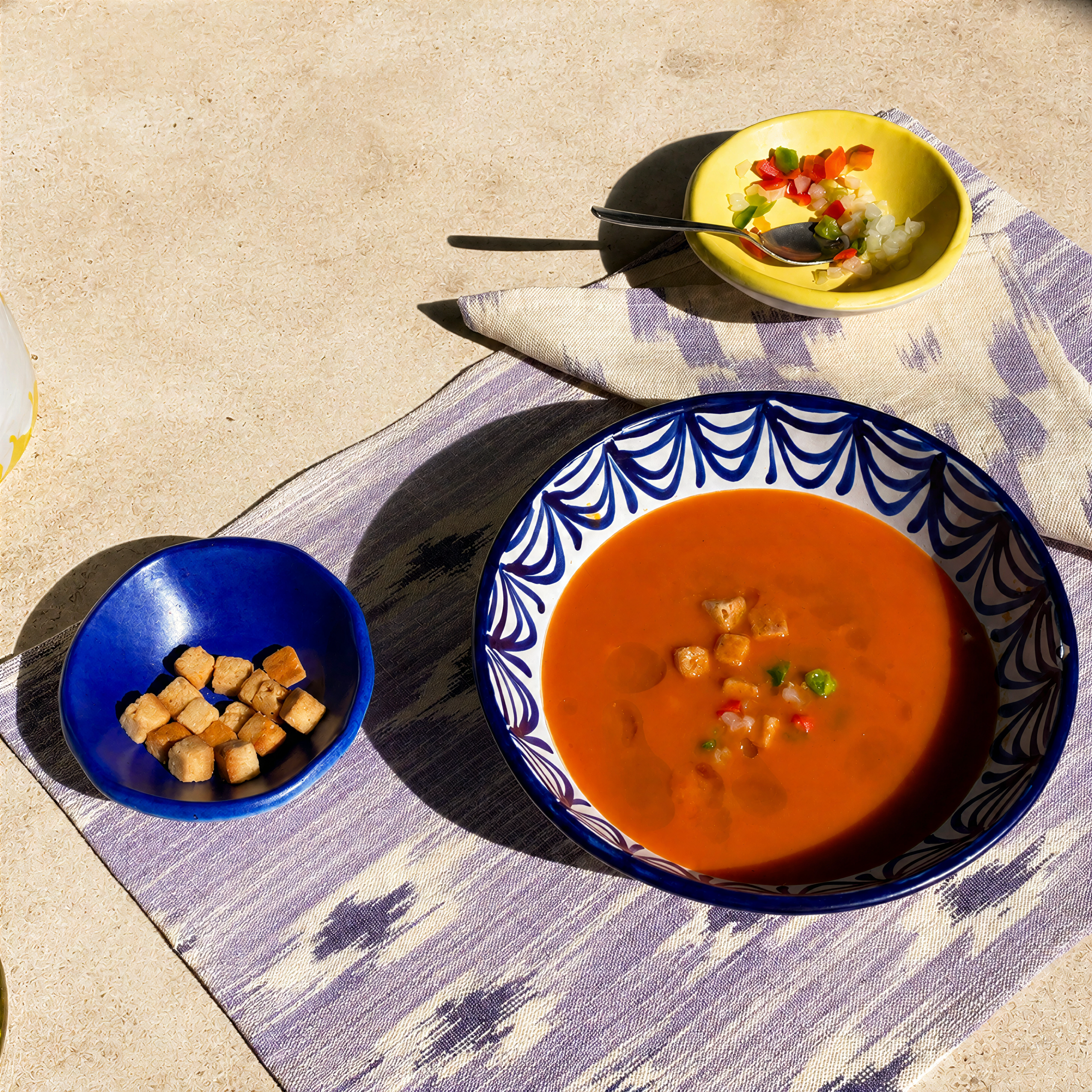 A styled outdoor dining scene featuring a large bowl of gazpacho with croutons in a blue and white patterned ceramic bowl, accompanied by a small blue bowl of croutons and a yellow bowl of rice on a patterned placemat. - lifestyle