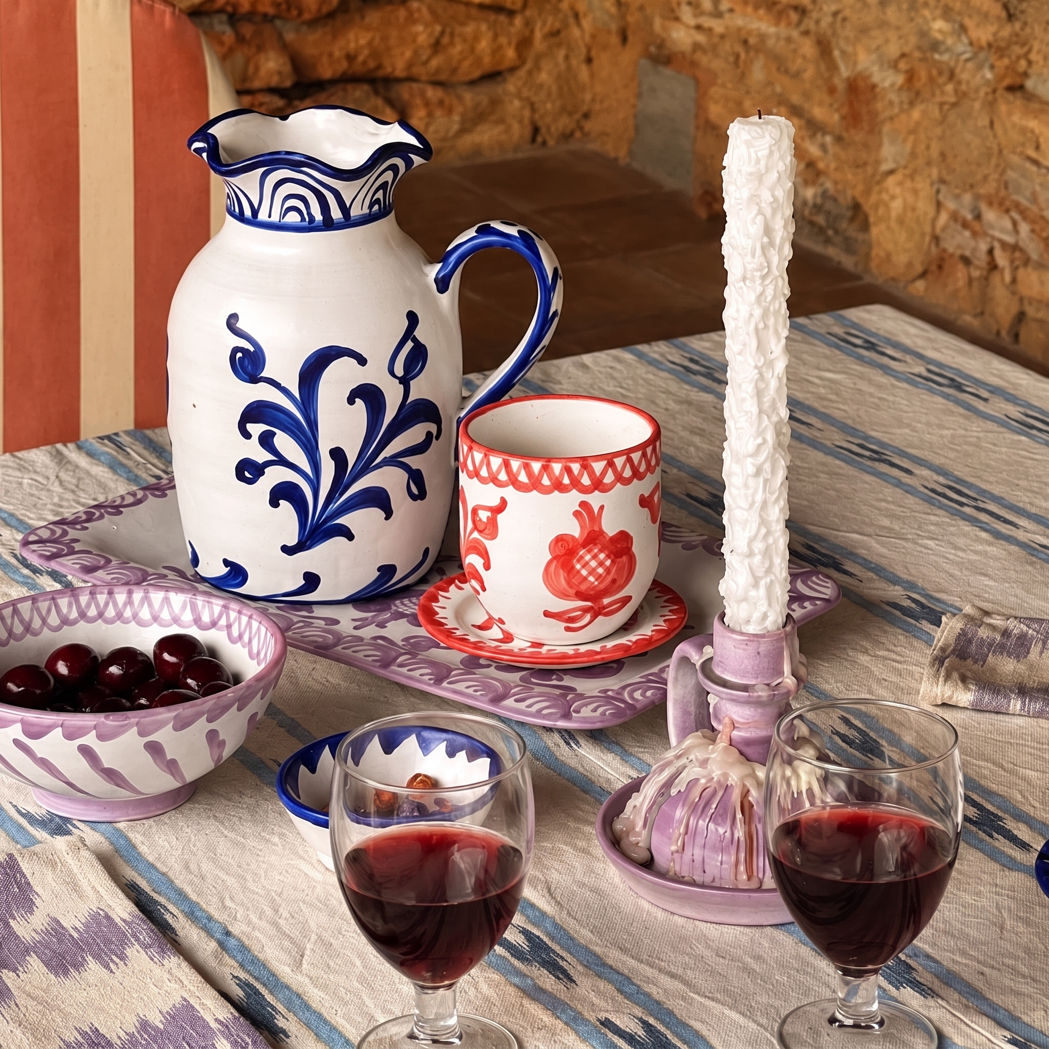 A styled table setting featuring a white ceramic pitcher and cup set with blue and red floral patterns, alongside glasses of red wine, a bowl of cherries, garlic, and a tall white candle on a patterned tablecloth. - lifestyle