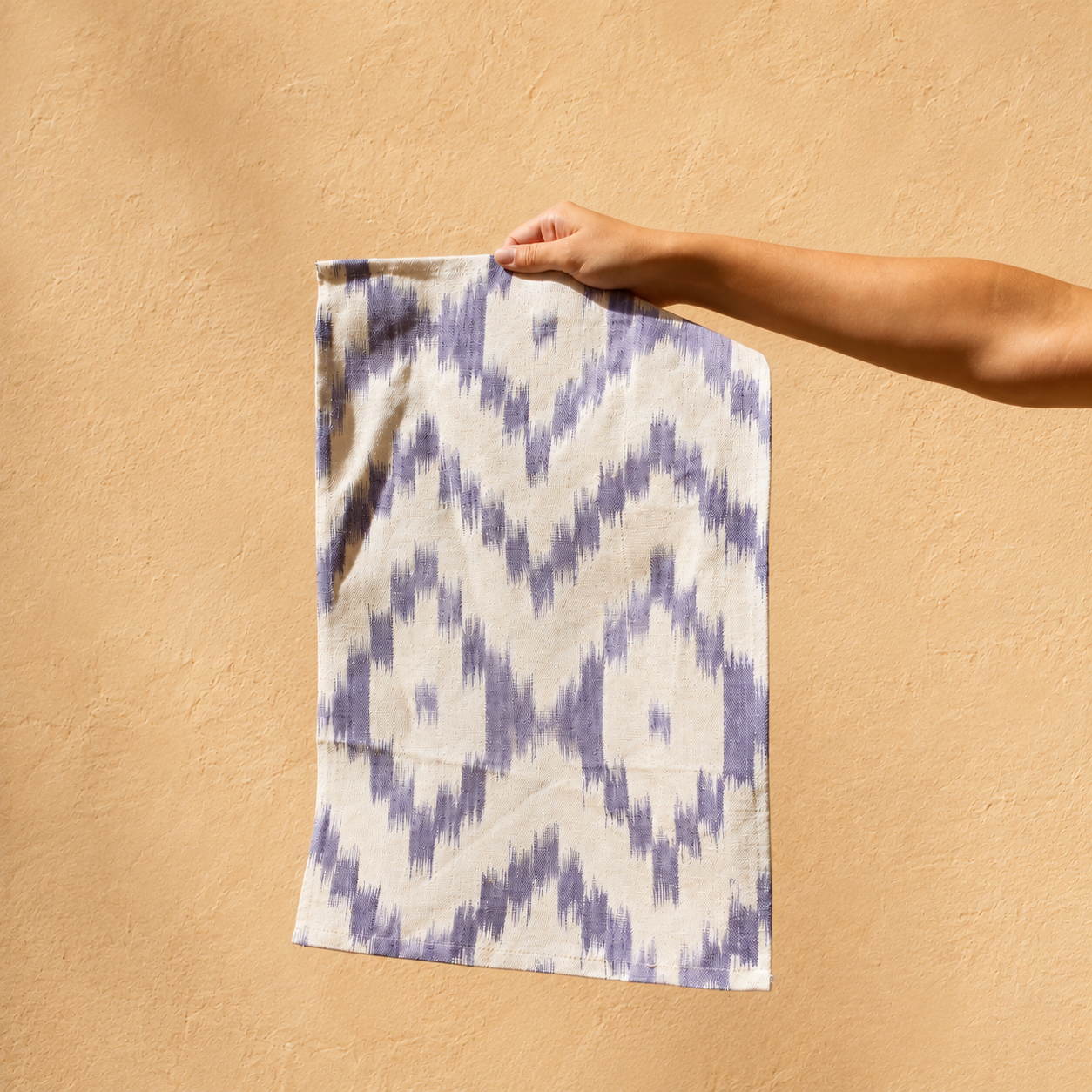A hand holds up a rectangular textile with a purple and off-white ikat-style geometric pattern against a warm beige stucco wall. - lifestyle