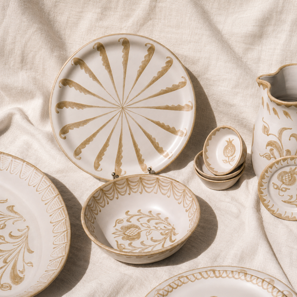 A coordinated set of white ceramic tableware with warm beige botanical and geometric patterns, arranged on draped linen fabric. Visible pieces include plates, bowls, a pitcher, and stacked small bowls. - lifestyle