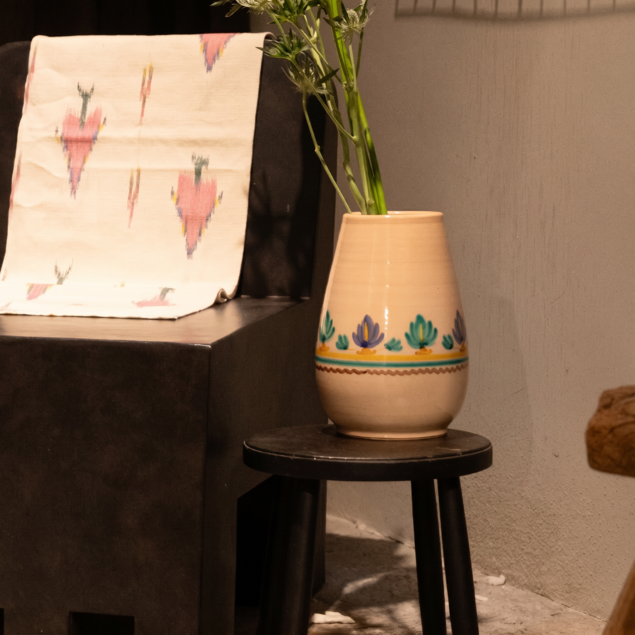 A beige ceramic vase with colorful hand-painted floral patterns sits on a small black stool, holding green bamboo stems. The scene is styled in a rustic indoor setting with a dark table holding a patterned cream cloth in the background. - lifestyle