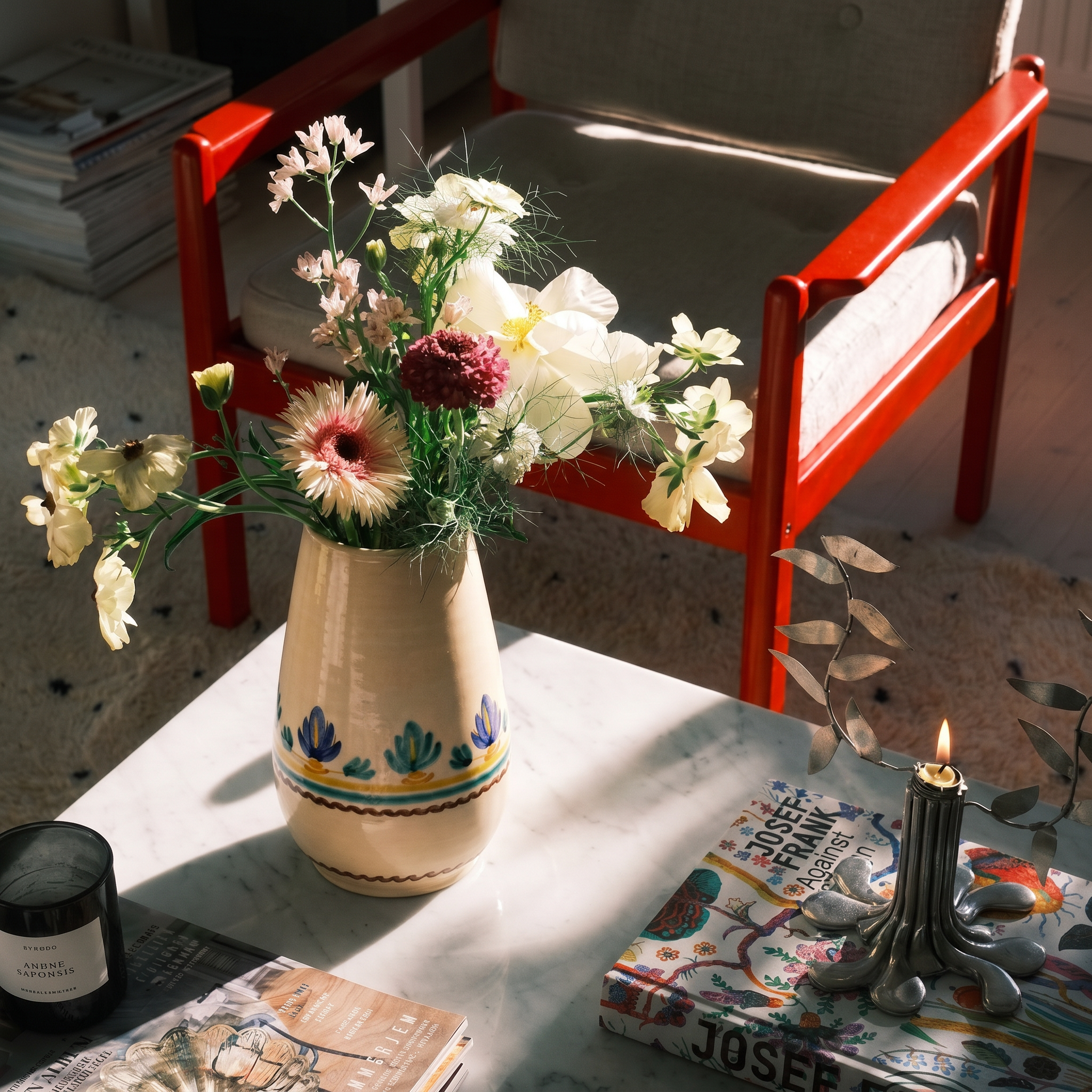 A cream ceramic vase with blue and green floral patterns holds a bouquet of white, pink, and burgundy flowers on a white marble table. A red wooden chair sits behind the table in warm sunlight, alongside books, a candle, and other home decor. - lifestyle