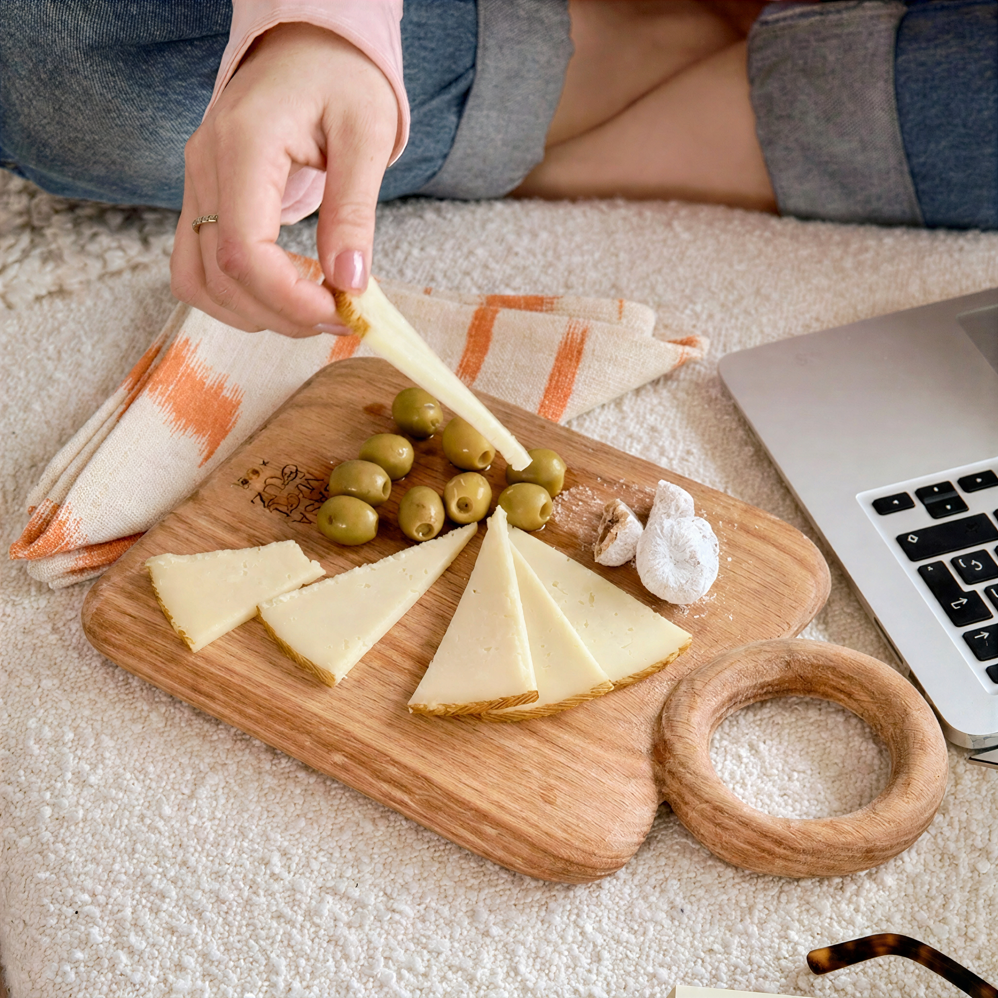 A wooden cheese board with sliced cheese, green olives, garlic, and a small ring-shaped wooden item, styled on a cream textured blanket with a person's hand placing cheese, a laptop, and a striped cloth nearby. - lifestyle