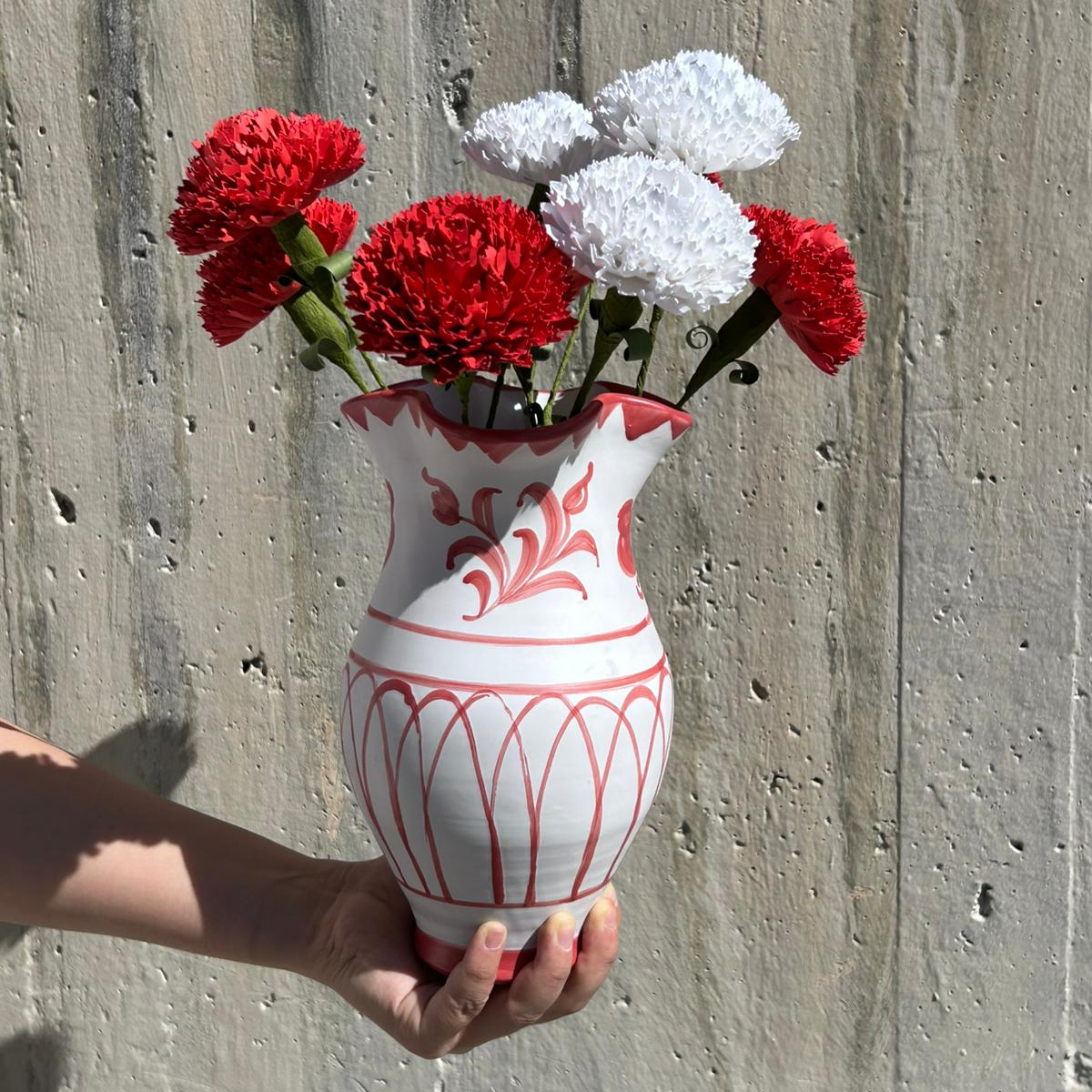 White vase with red floral patterns holding red and white flowers against a concrete wall.