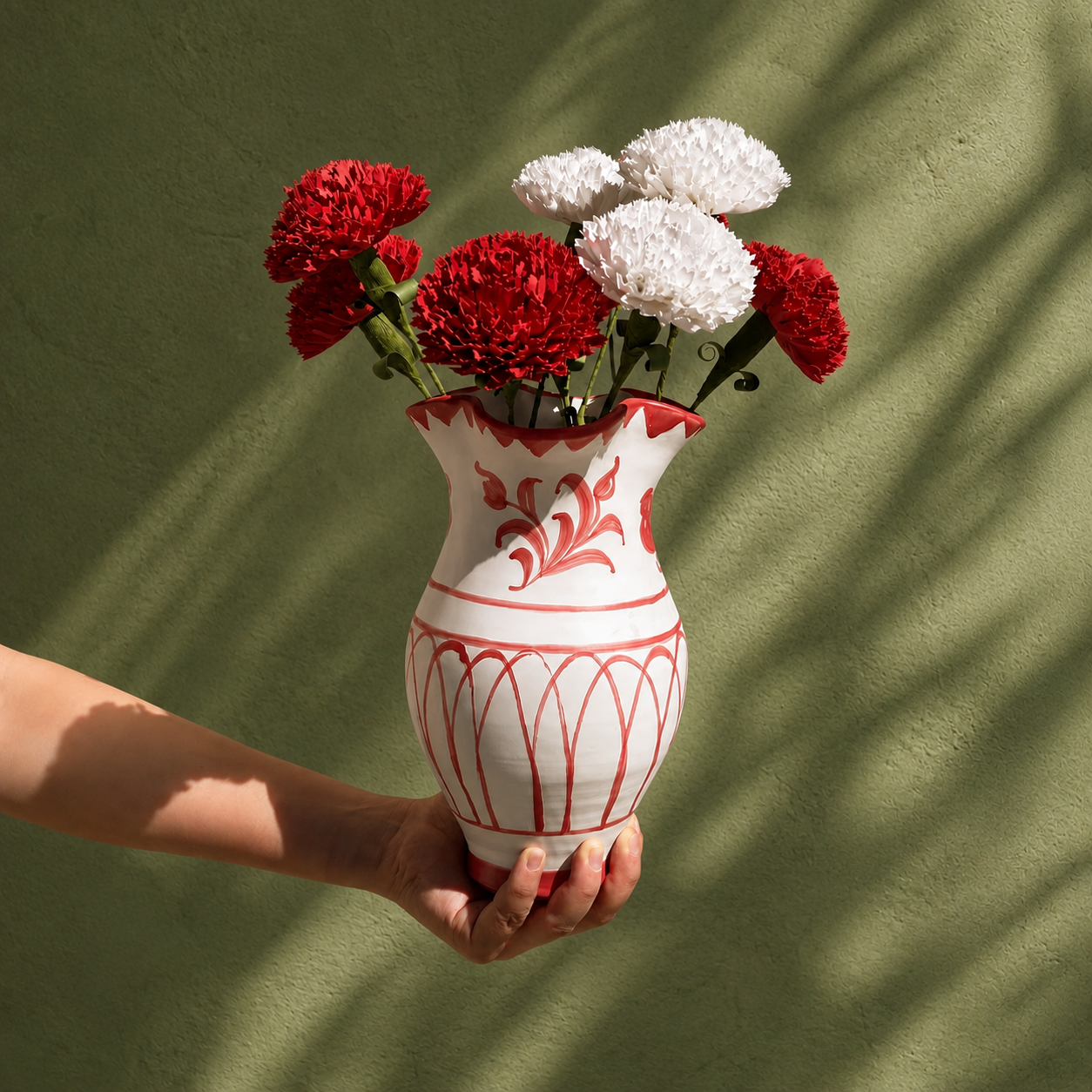 A hand holds a white ceramic vase with red painted patterns, filled with red and white flowers, against a textured olive green wall with soft leaf shadows. - lifestyle