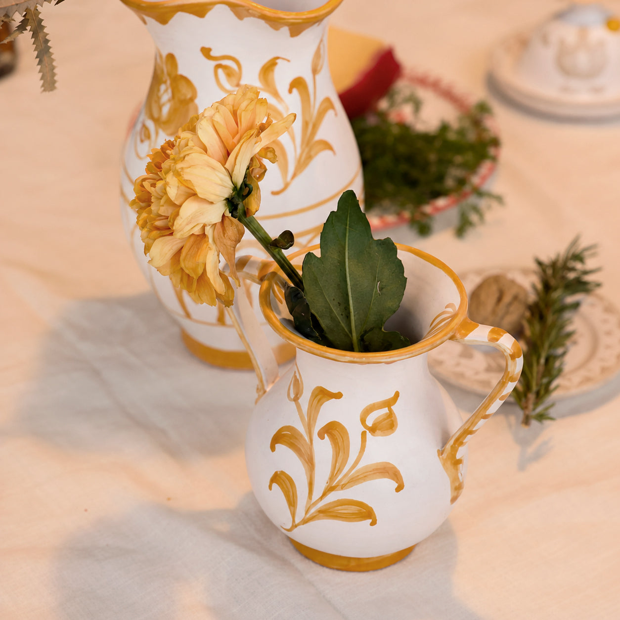 Two white ceramic pitchers with gold floral scrollwork and handles, styled on a light fabric surface with a yellow flower in the front pitcher and another pitcher behind it. - lifestyle