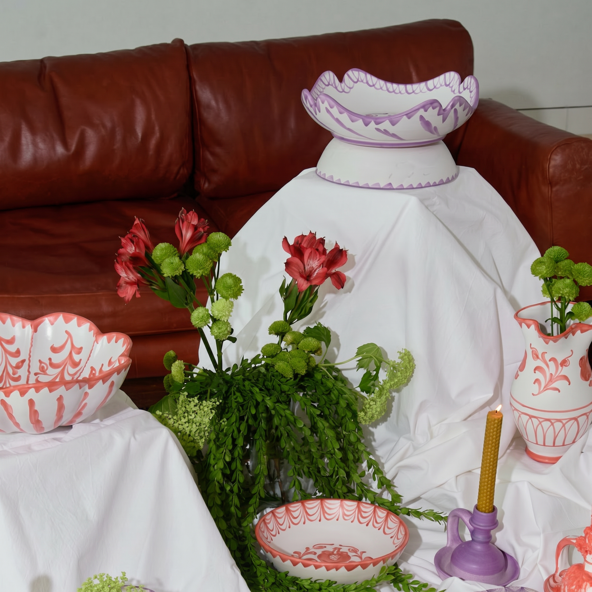 A collection of white ceramic tableware with coral-red patterns, including bowls, a vase, and a candle holder, styled on a white tablecloth in front of a brown leather sofa. - lifestyle