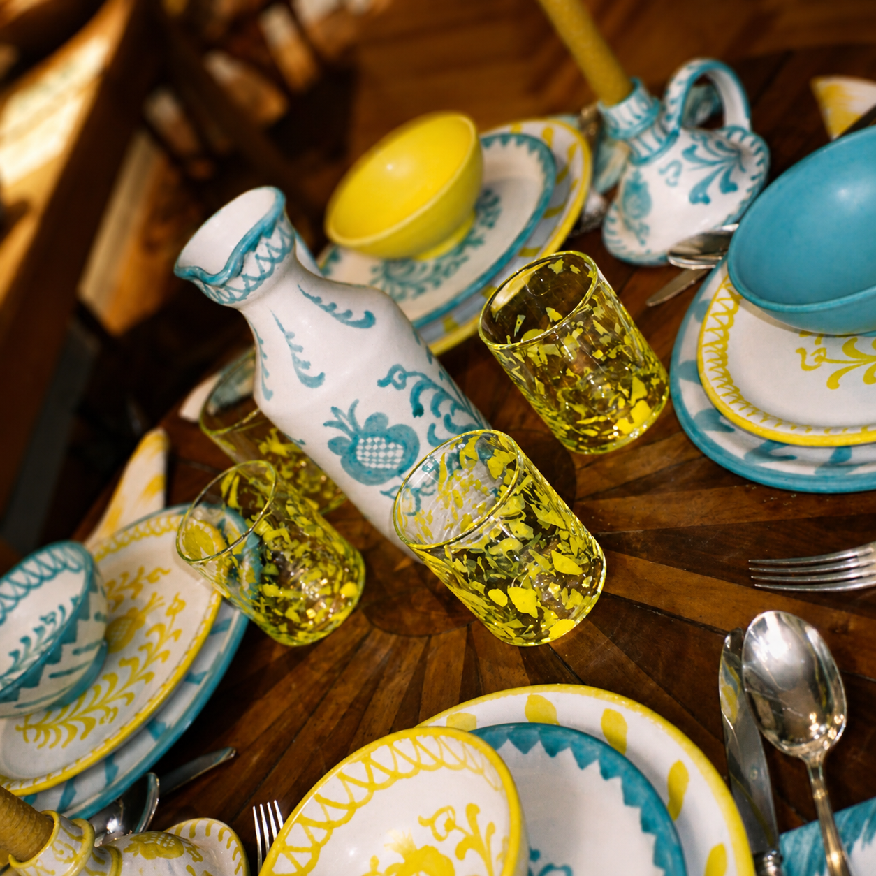 A styled dining table setting featuring a white ceramic carafe and plates decorated with teal and yellow patterns, paired with yellow and teal glass tumblers, set on a dark wooden table. - lifestyle