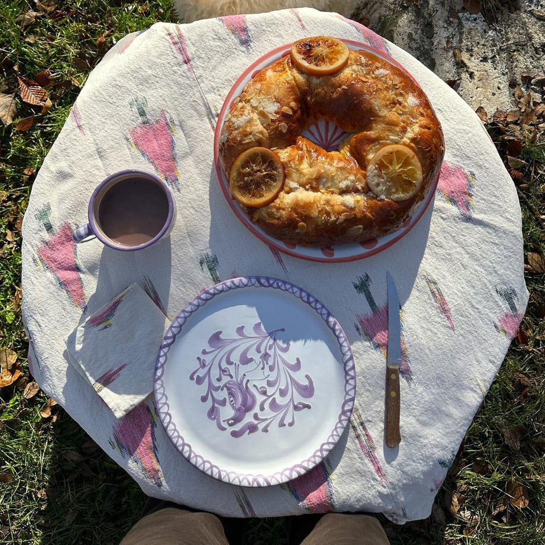 Round table with a cake, traditional dinner plate, linen, and a cup on a grassy surface with a dog nearby.