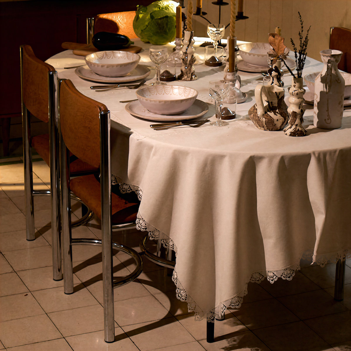 An off-white lace-trimmed round tablecloth draped over a set dining table with white plates, glassware, and wooden chairs in a restaurant setting. - lifestyle