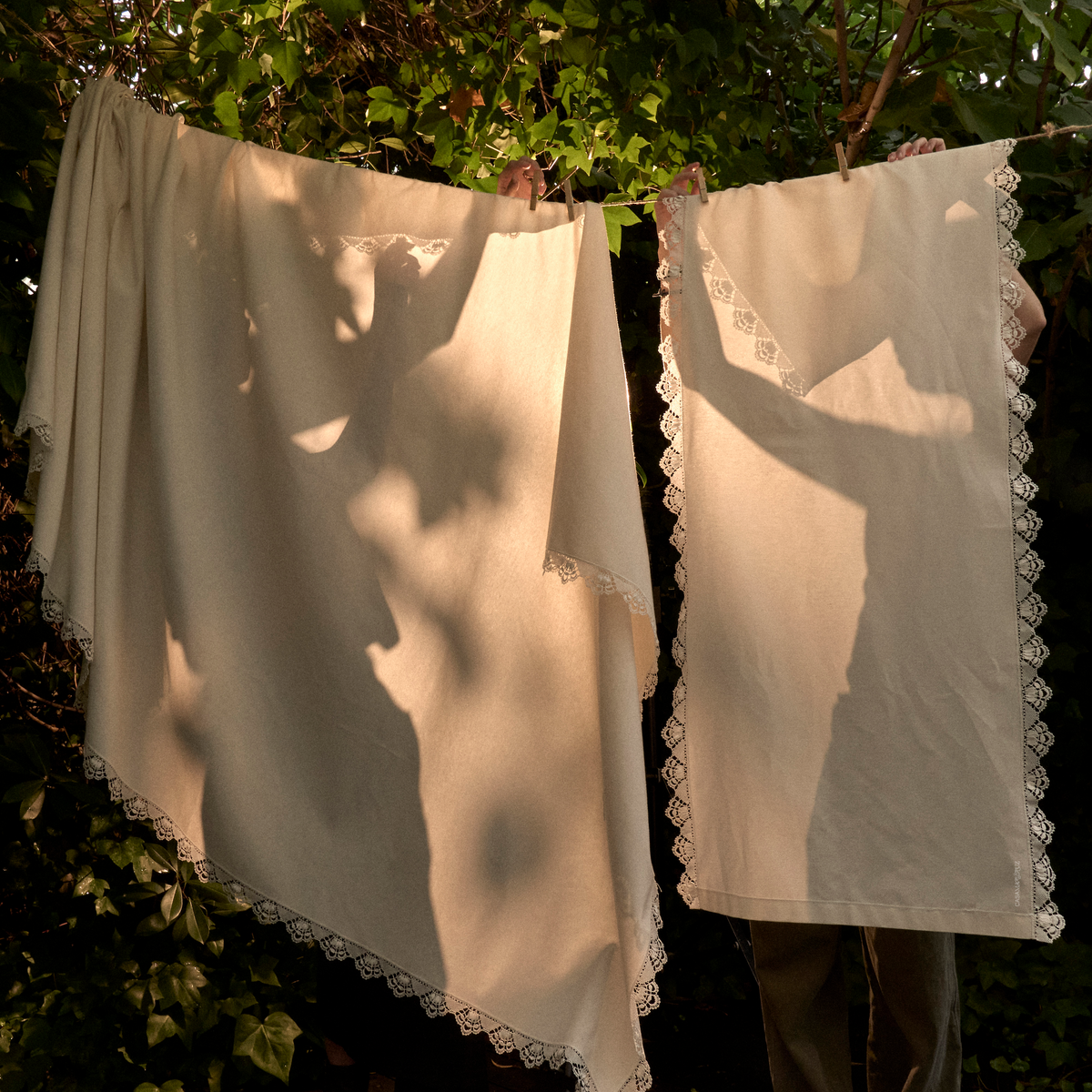Two cream-colored linen cloths with delicate lace trim hanging outdoors on a line against a backdrop of green foliage and dappled sunlight. - lifestyle