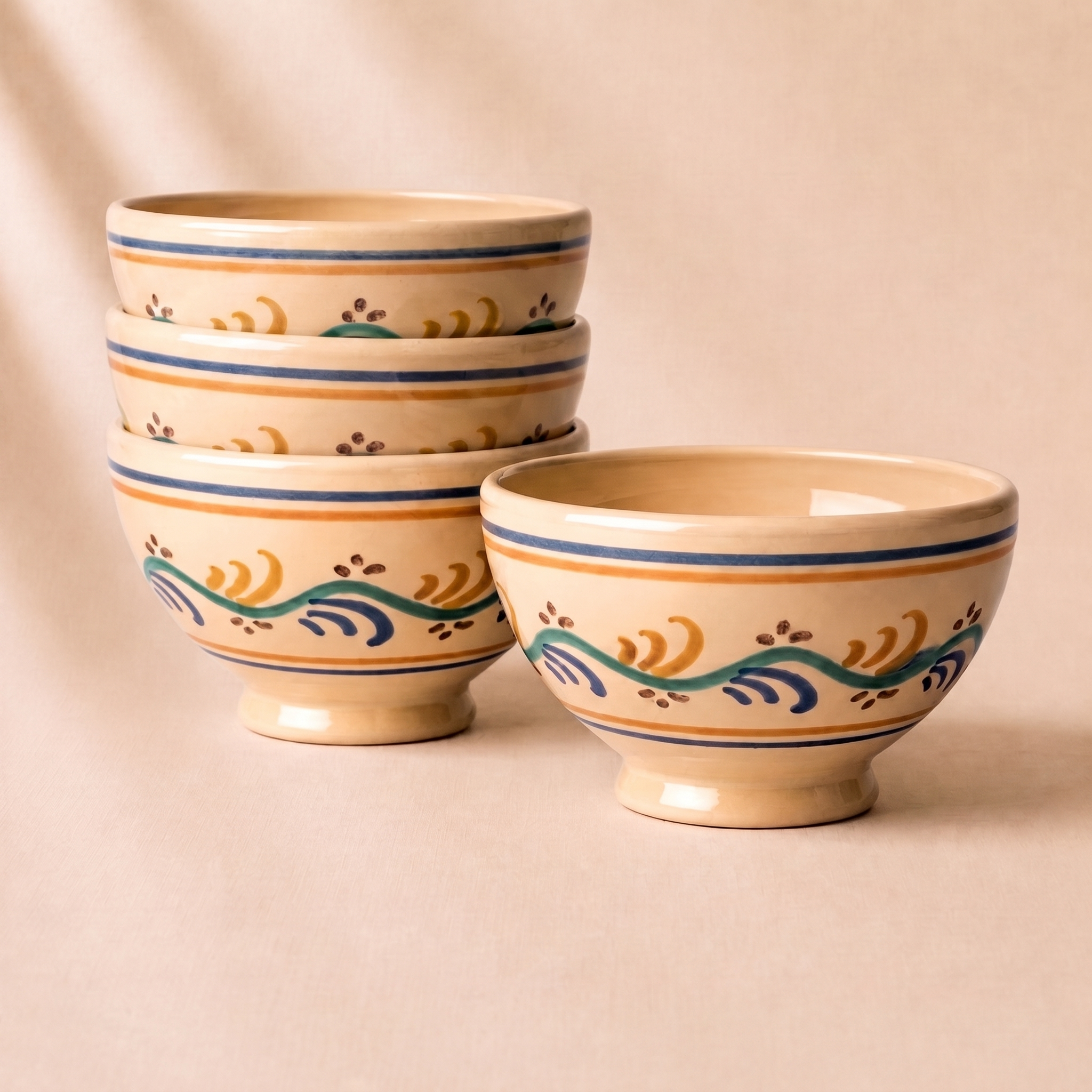 Three beige ceramic bowls with blue, green, and orange hand-painted wave patterns, with two stacked on top of each other and one standing in front, set against a warm beige background.