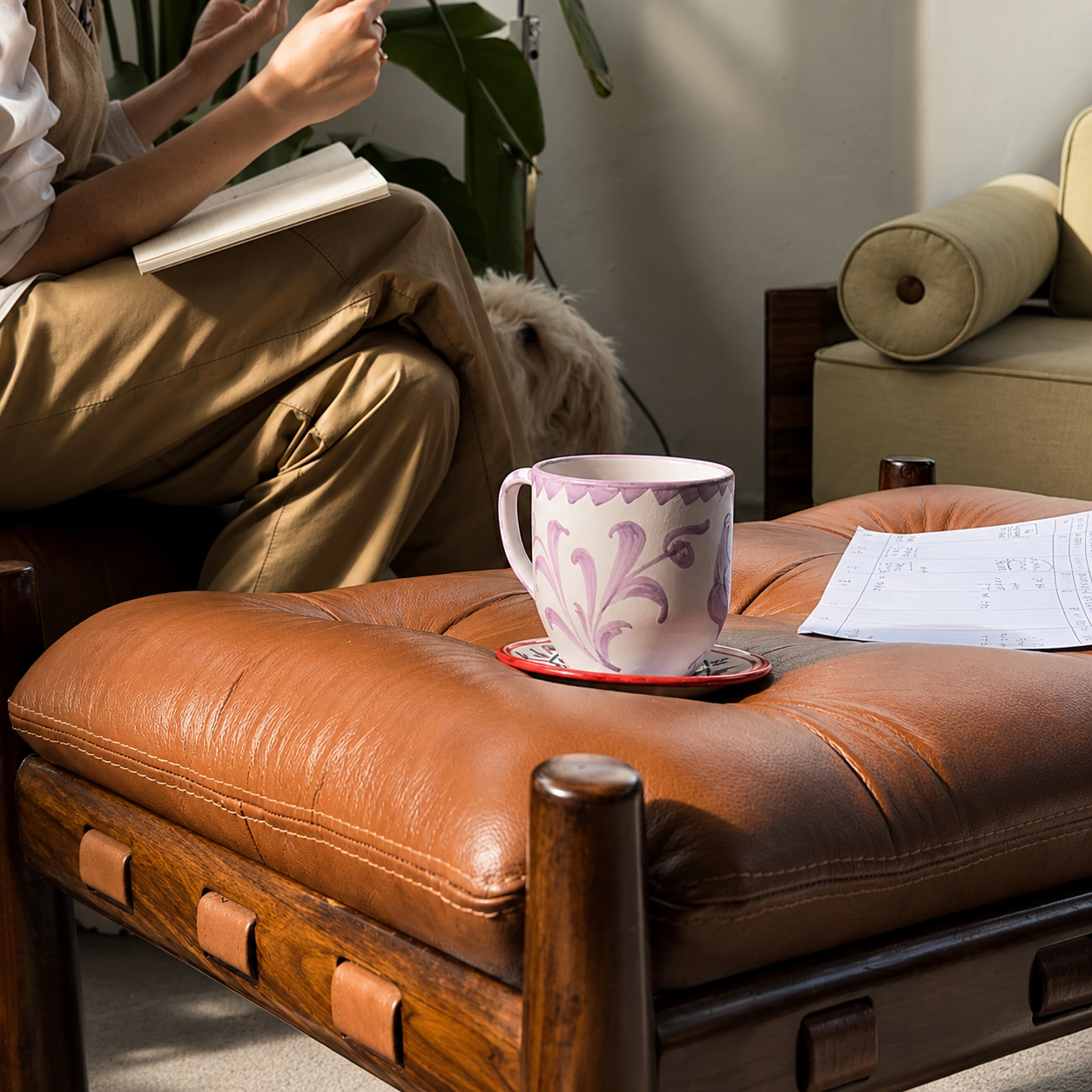 A brown leather ottoman with a dark wood frame in a cozy living room setting, with a pink floral-patterned mug on a saucer resting on top. - lifestyle