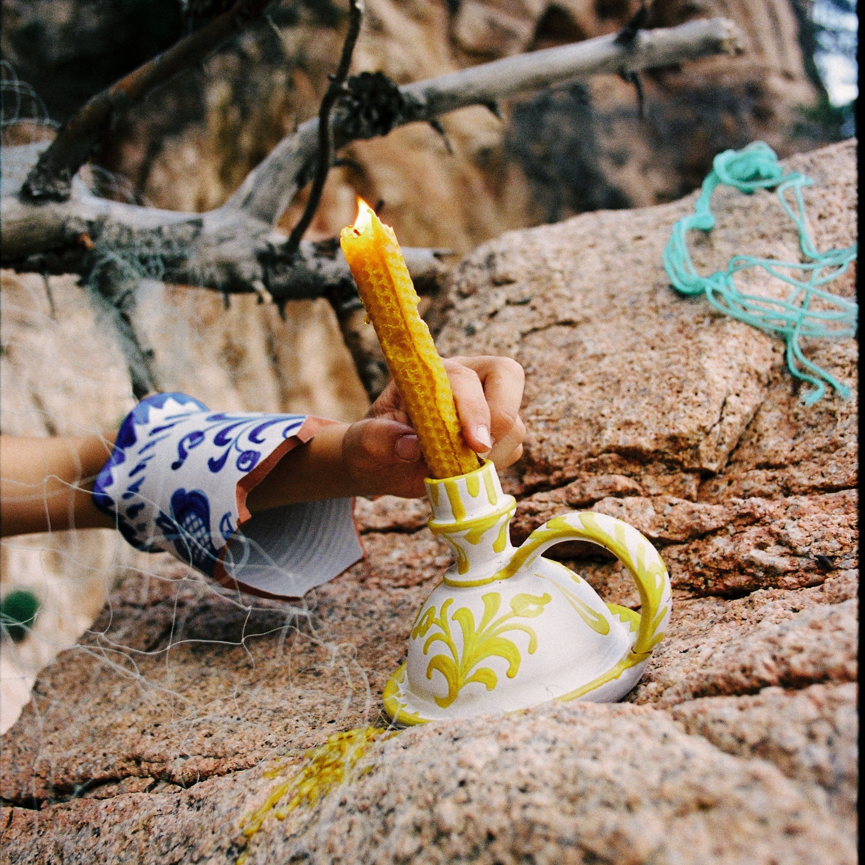 Hand holding a lit candle inside a decorative ceramic holder on a rocky surface with natural background