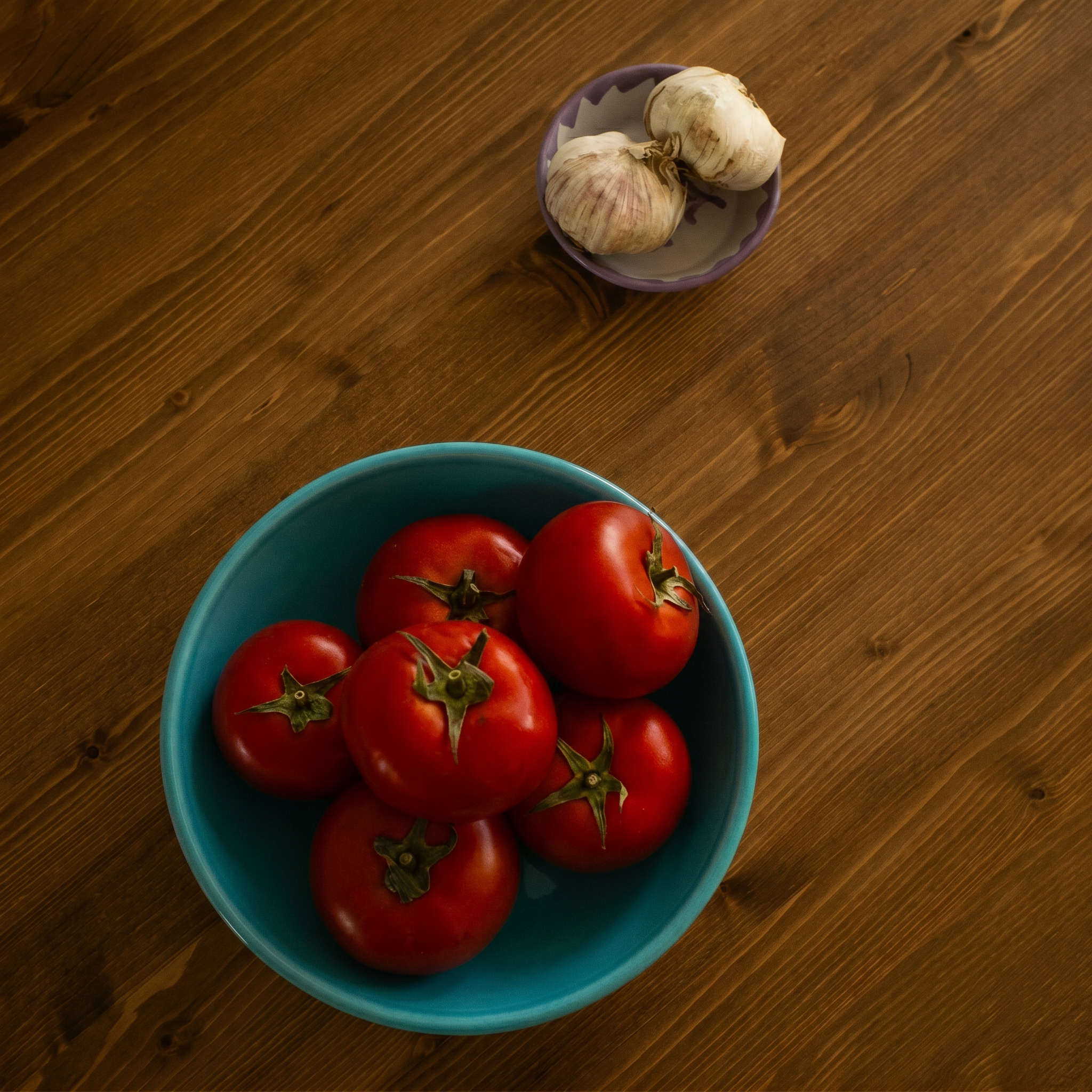 A turquoise bowl filled with red tomatoes on a wooden table, with a small purple dish of garlic in the background. - lifestyle