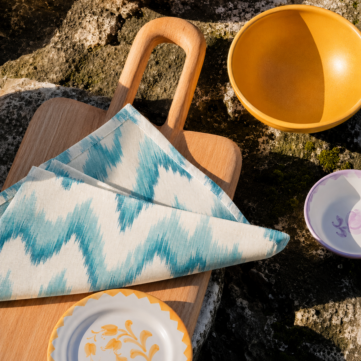 A folded patterned cloth napkin with a blue and white ikat design rests on a wooden cutting board outdoors on a rocky surface. A yellow bowl and a small white plate with yellow floral detailing are also visible in the scene. - lifestyle