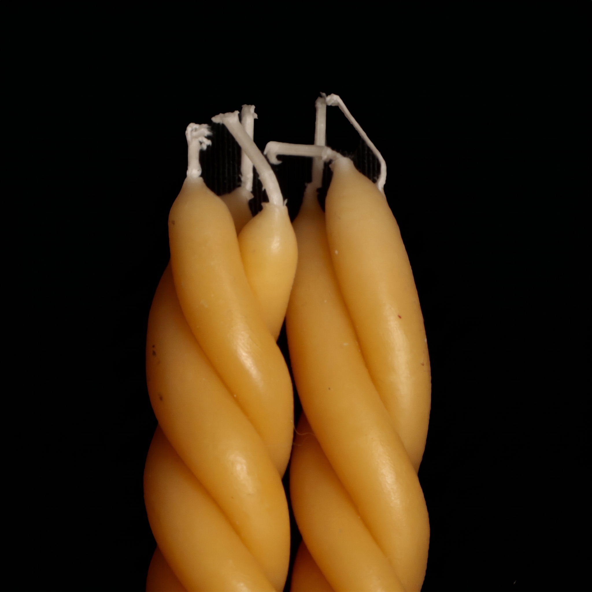 Two twisted taper candles in a warm pale yellow wax, standing upright against a solid black background. - lifestyle