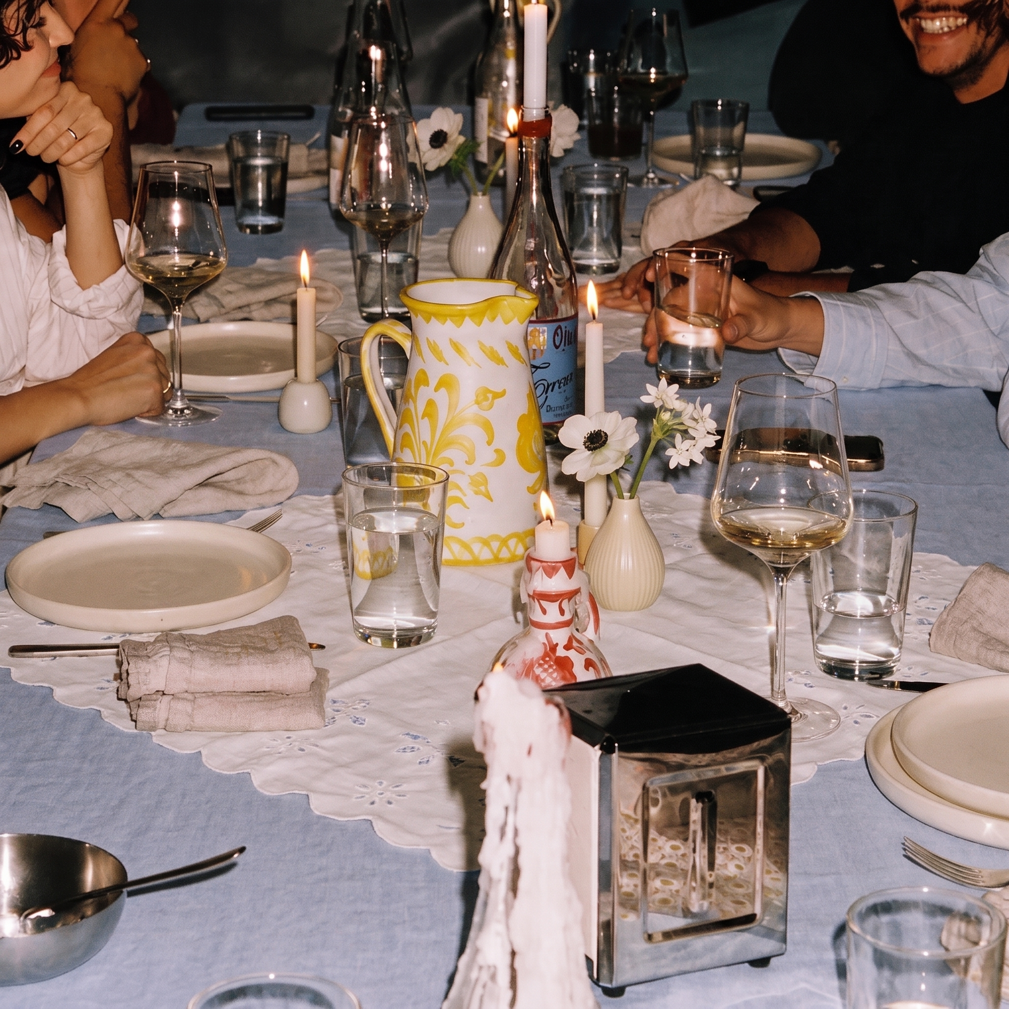 A cozy, dimly lit dinner table scene with people gathered around, holding glasses and sharing a meal. The table is decorated with a white lace runner, a yellow and white patterned ceramic pitcher, candles, wine glasses, and plates. - lifestyle