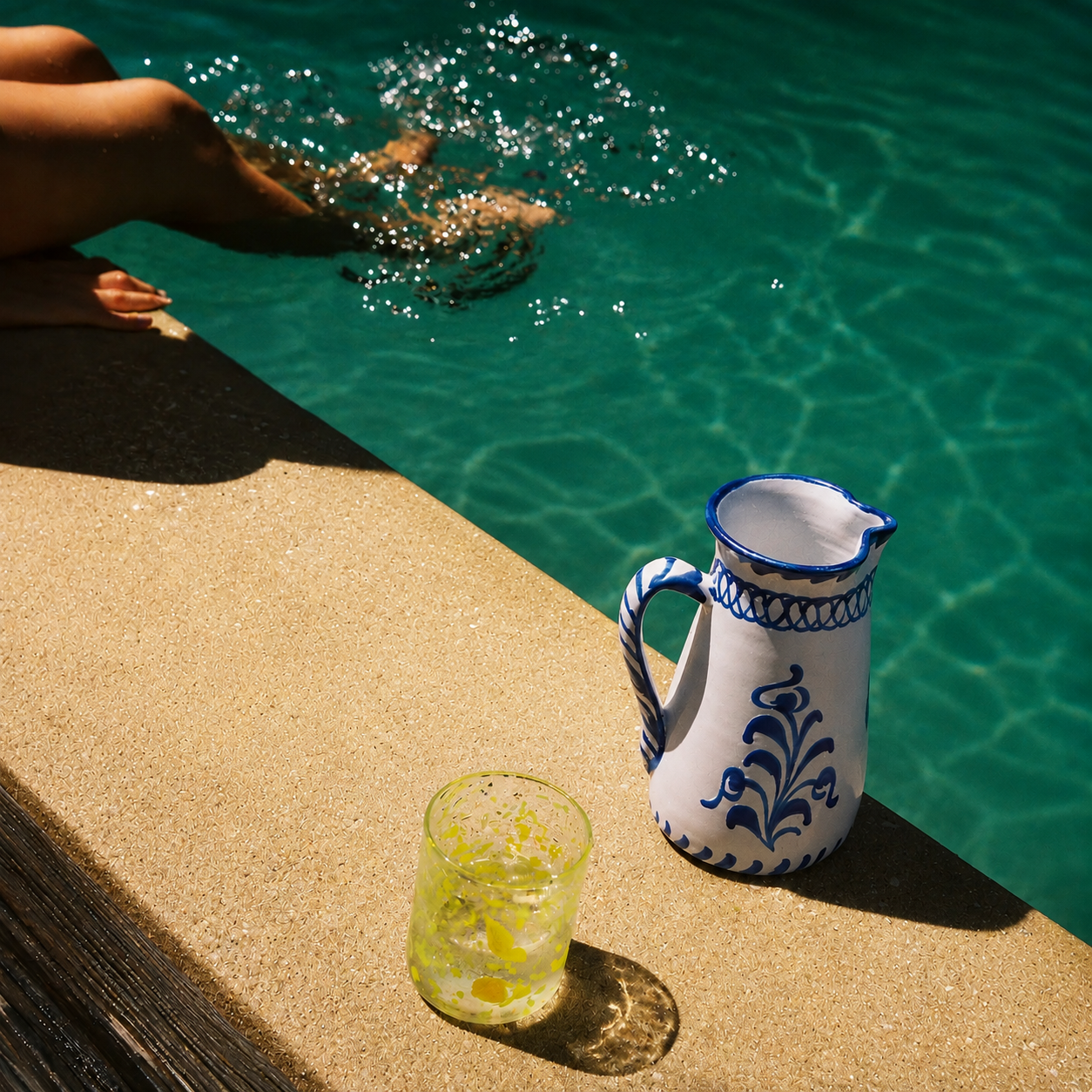 A white ceramic pitcher with blue floral patterns sits on a concrete poolside ledge next to a glass of iced lemonade, with a person's legs and feet visible in the turquoise swimming pool water in the background. - lifestyle