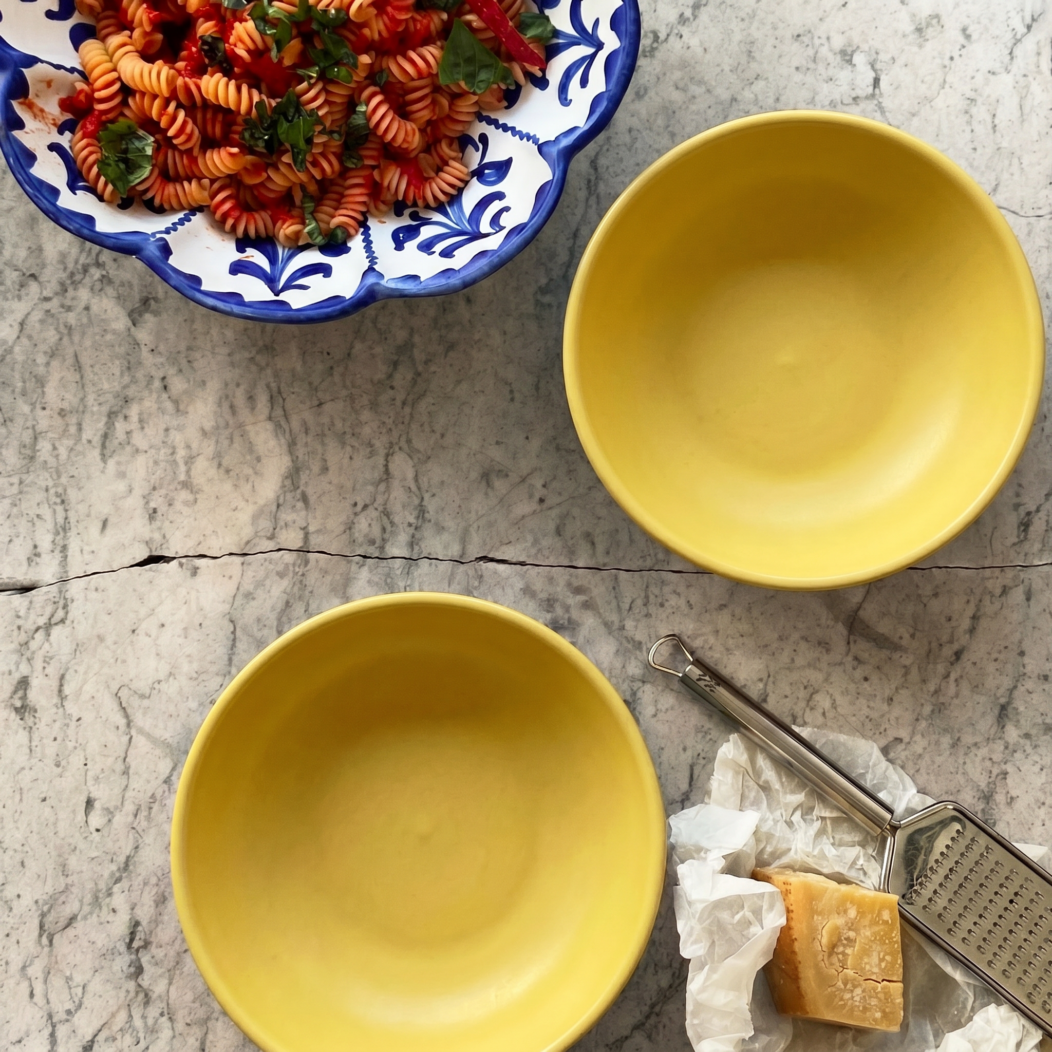 Two yellow ceramic bowls on a gray marble surface, with a bowl of pasta in the top left and a grater with cheese in the bottom right. - lifestyle